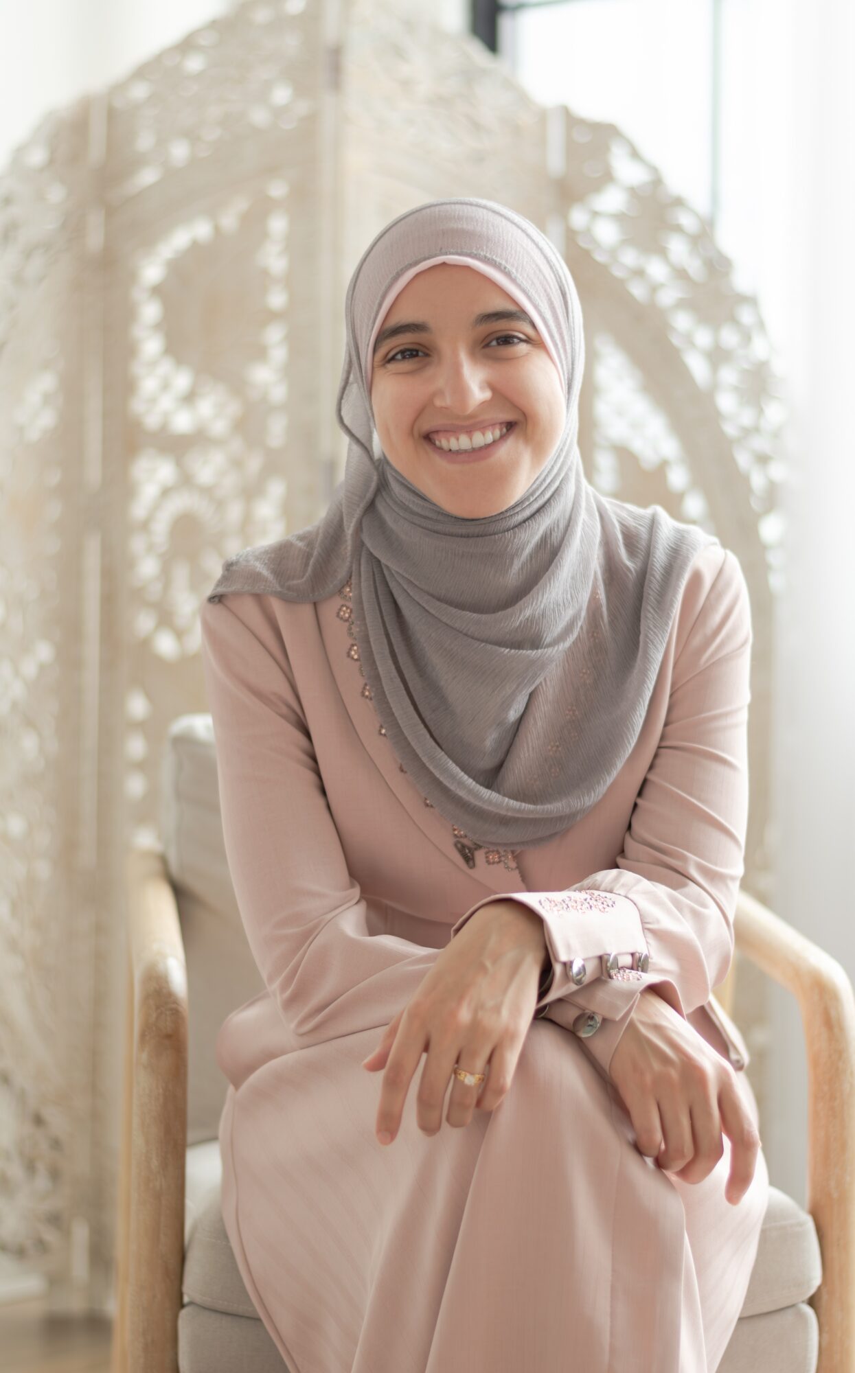 Smiling woman wearing a hijab and modest clothing, sitting indoors with decorative background elements.