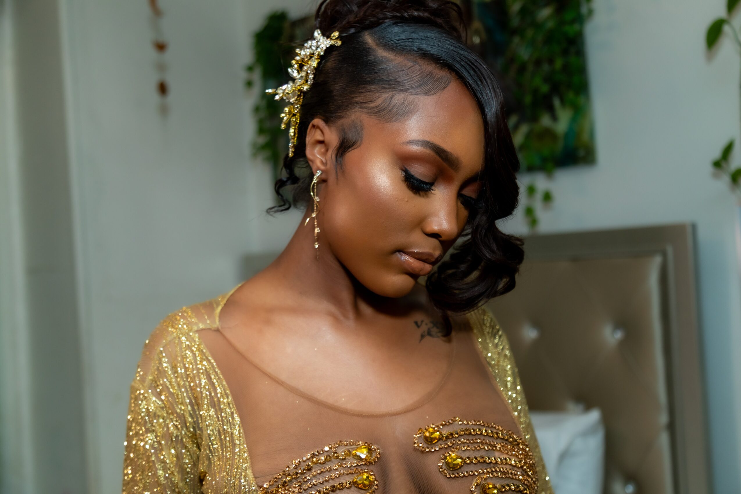 Woman with styled hair and gold accessories, wearing a gold dress, in a room with a bed and green plants