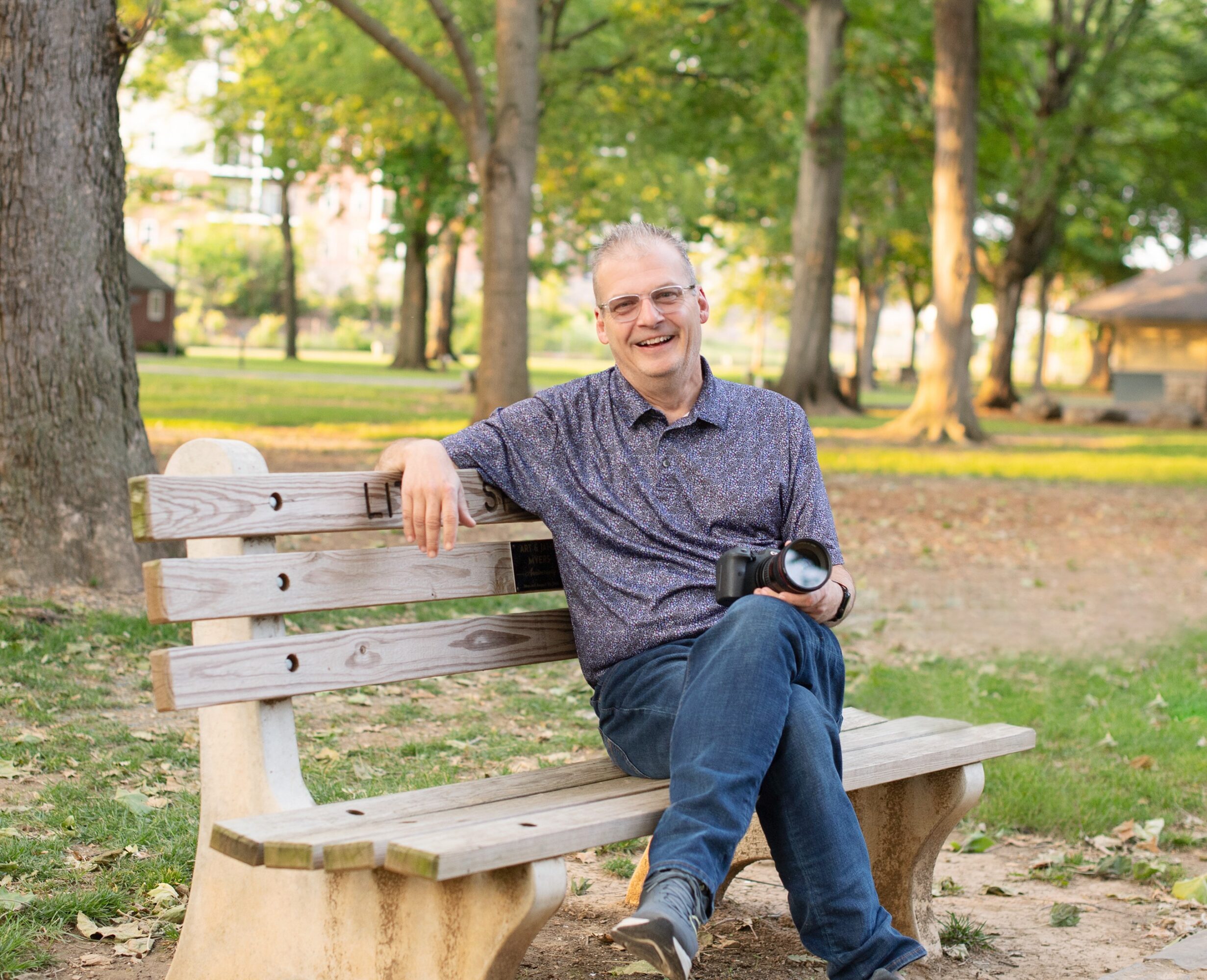 Man sitting on park bench smiling, holding a camera, surrounded by trees and greenery.