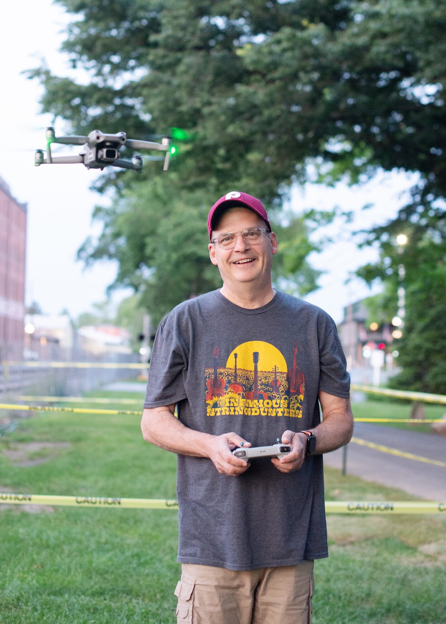 Person smiling outdoors holding a drone controller, with a drone flying nearby, trees and a park in the background.