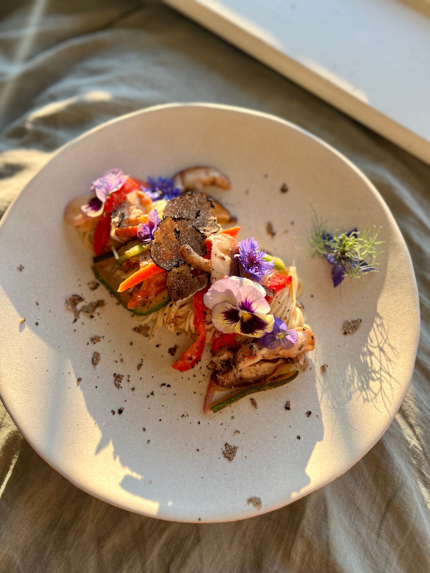 Plate with colorful food garnished with edible flowers and herbs, on a beige cloth surface, sunlight shining.