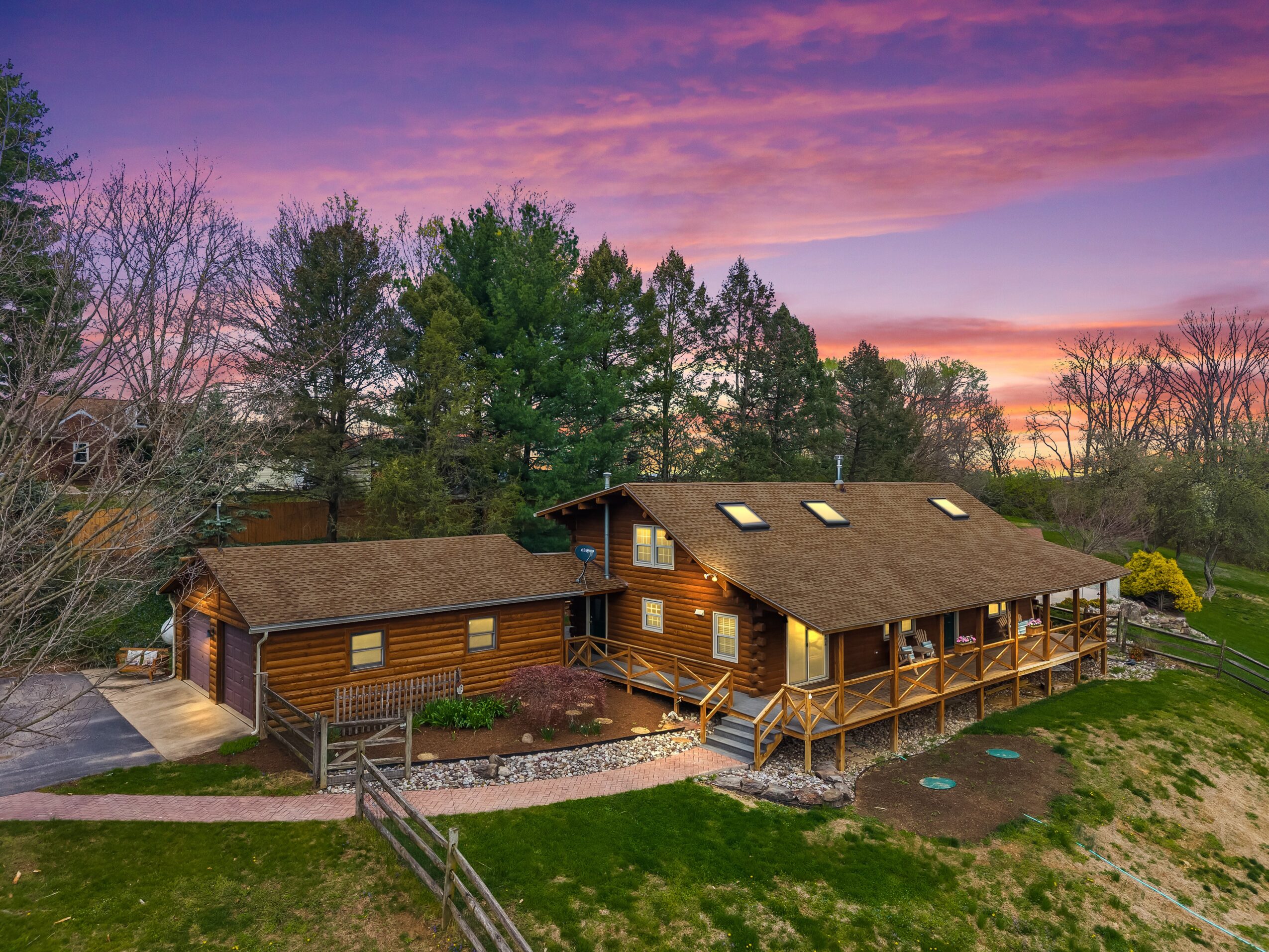 A wooden house with a deck, surrounded by trees and a grassy yard, under a colorful sunset sky.