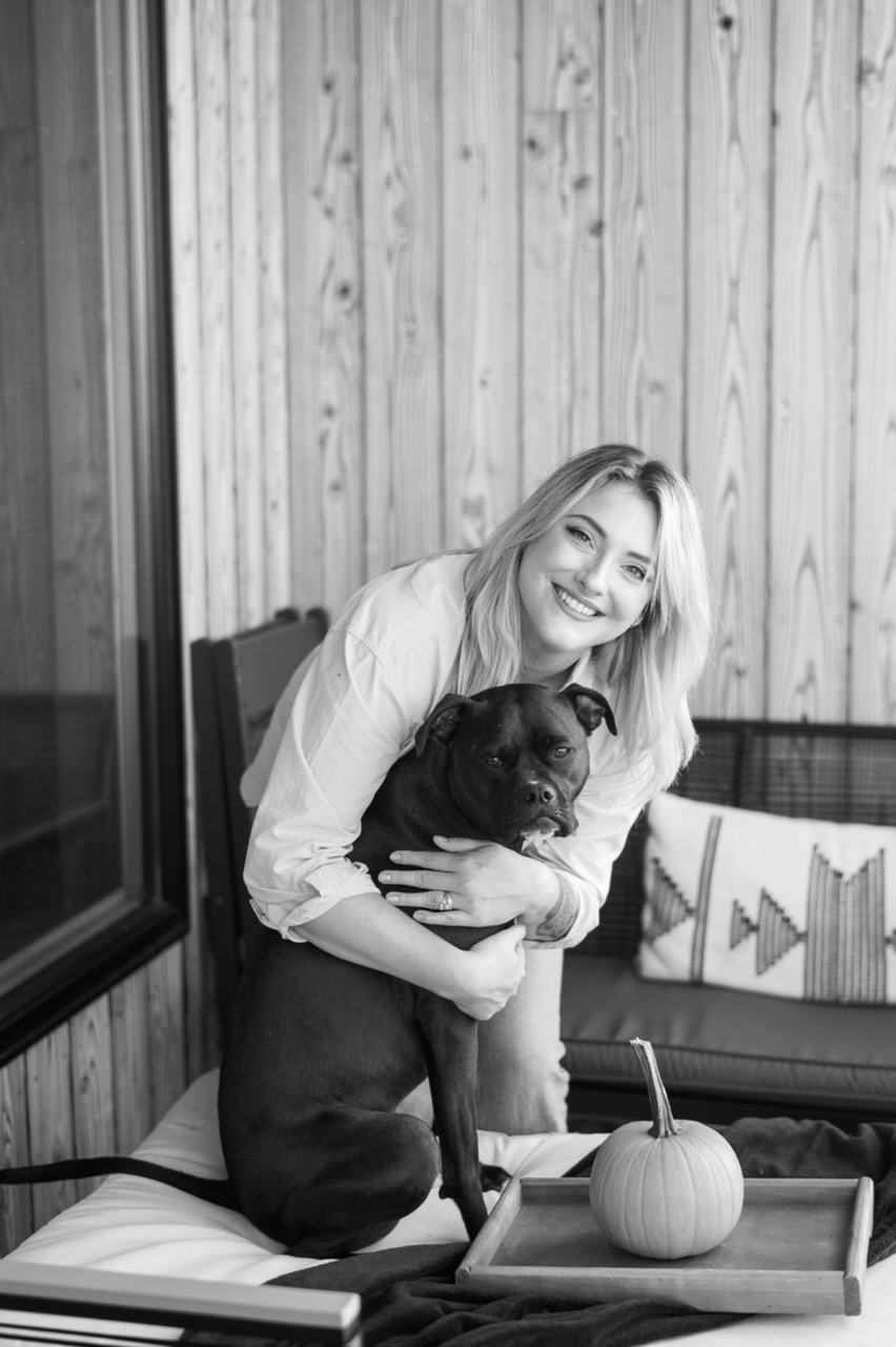 Woman smiling and hugging a black dog indoors with wooden wall background and decorative pillow.