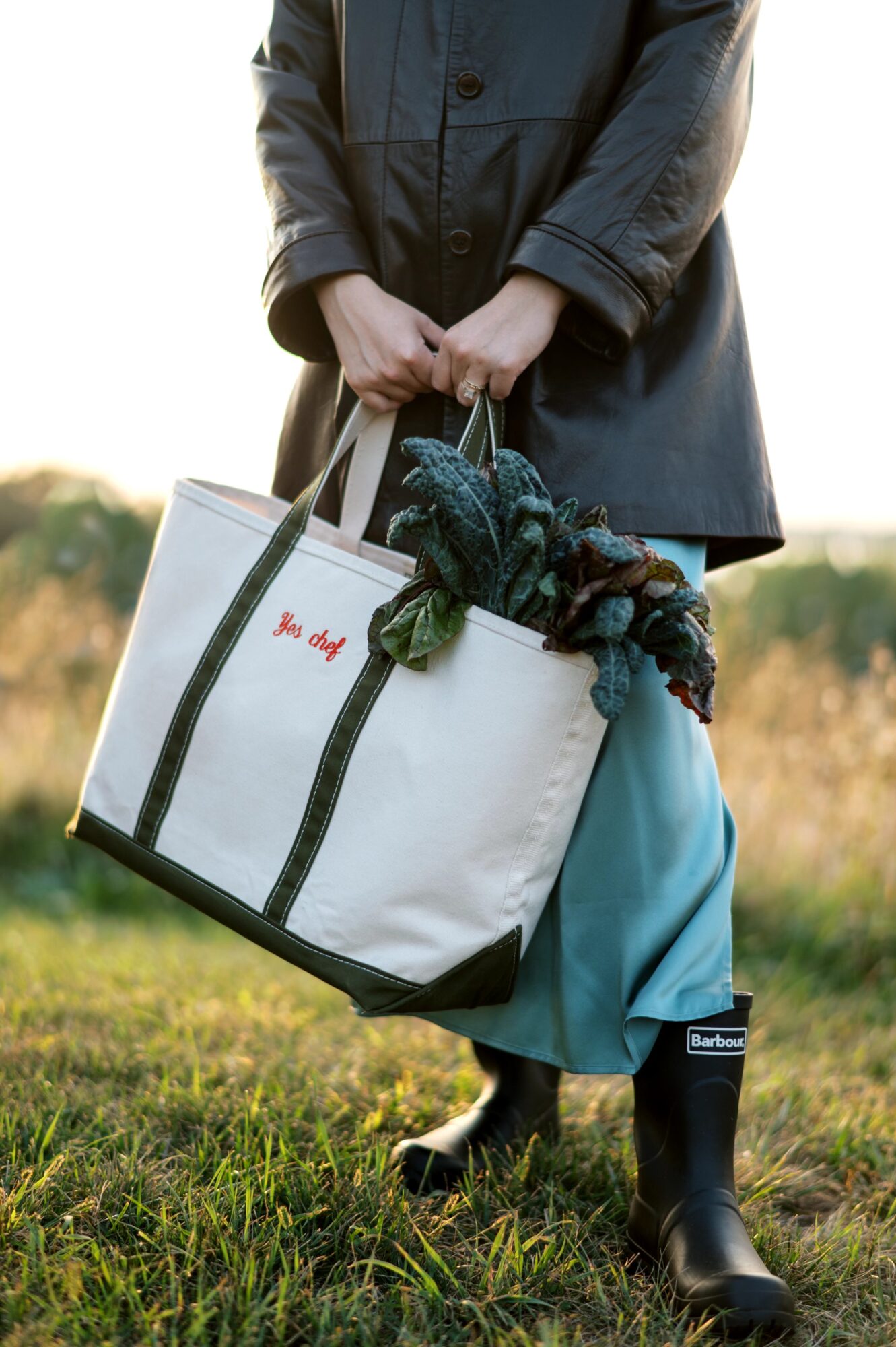 Person holding a large white bag with green handles, standing outdoors in a grassy field, wearing a dark coat and blue pants.