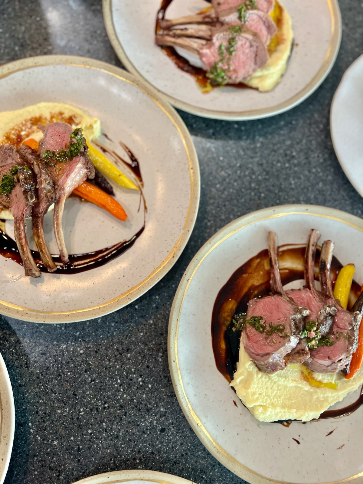 Three plates with lamb chops, vegetables, and sauce on a dark countertop, viewed from above.