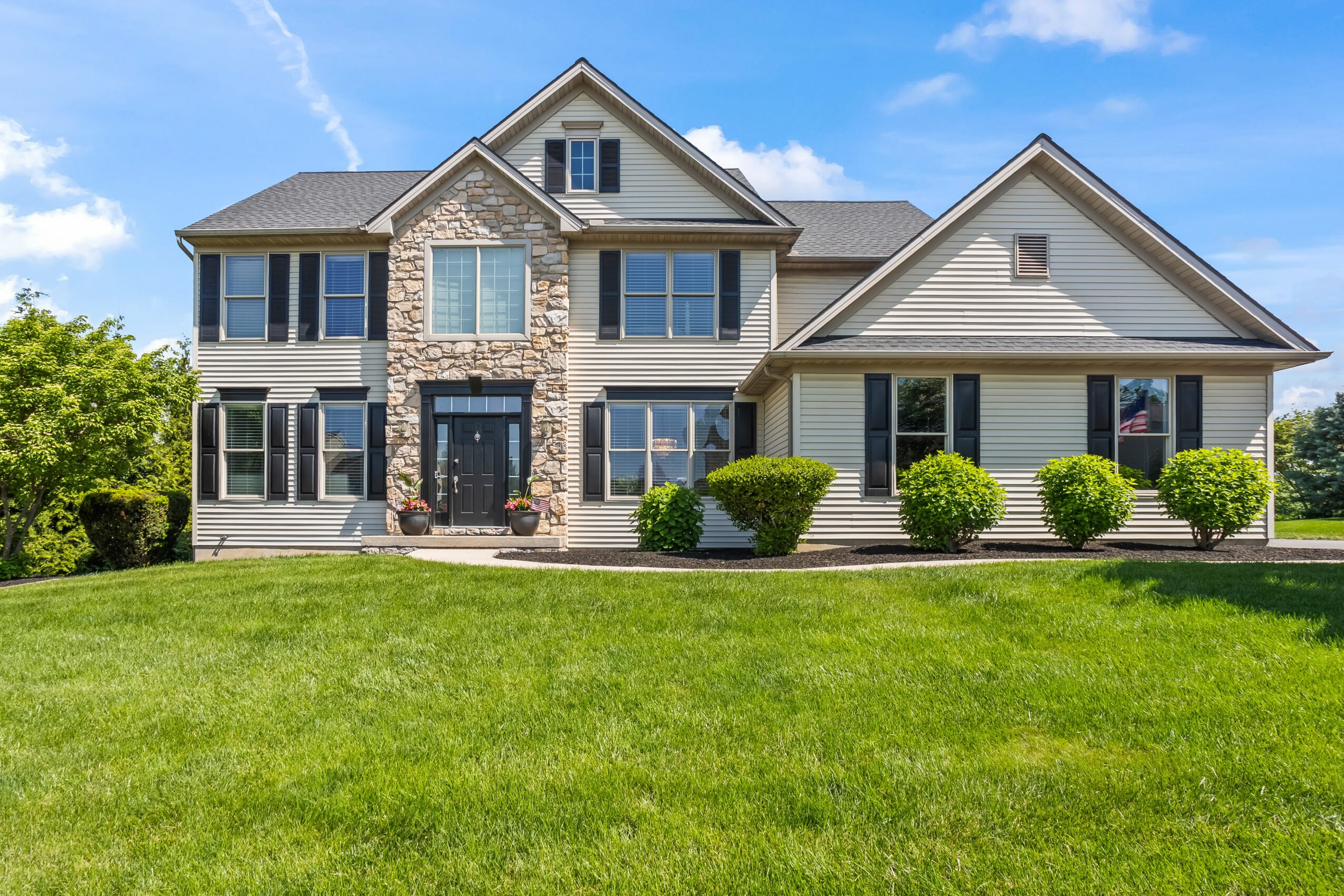 Large house with stone and siding exterior, multiple windows, and a well-maintained lawn with bushes in front.