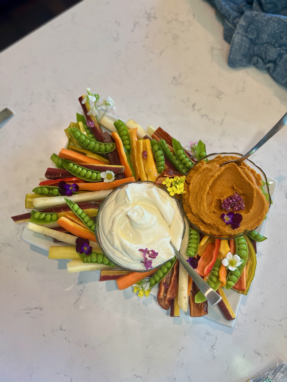 Colorful assortment of vegetables, dips, and a muffin on a white surface, with a person wearing blue jeans nearby.