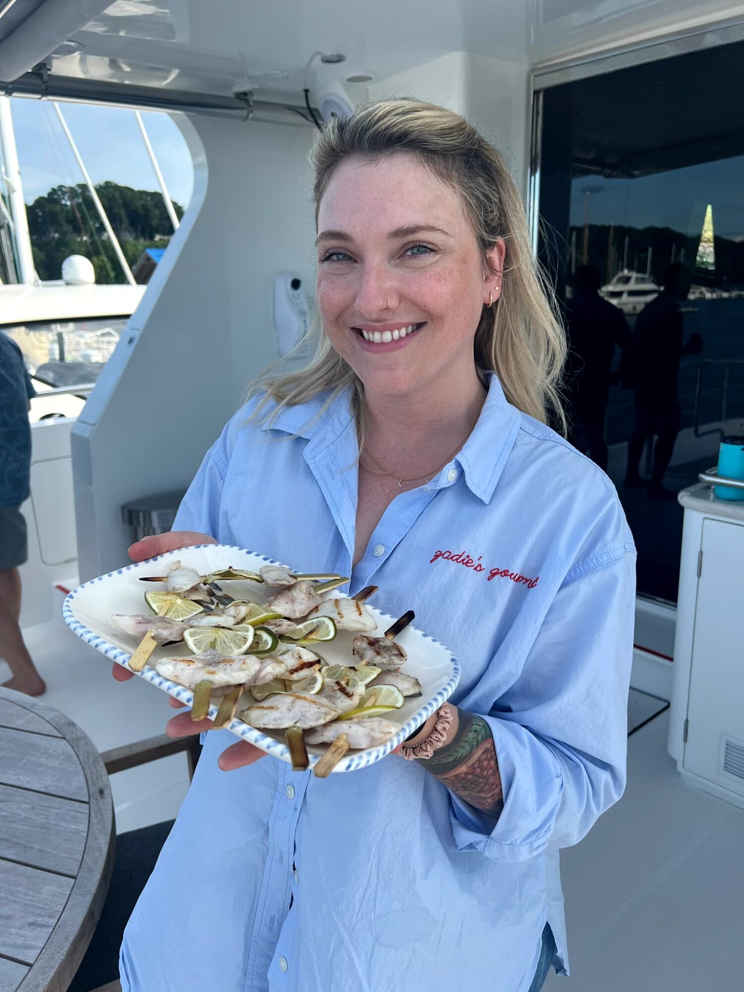 Woman with blonde hair smiling, holding a plate of oysters on a boat, wearing a light blue shirt.