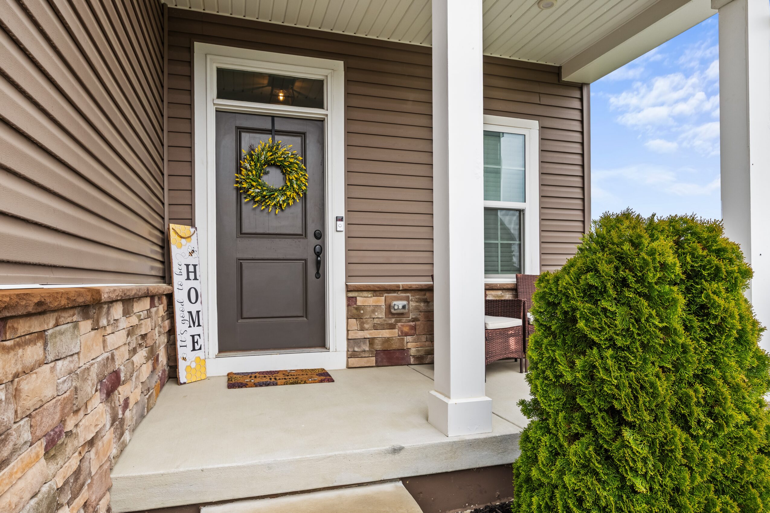 Front porch with door, wreath, and a large green bush, part of a house with siding and stone accents.