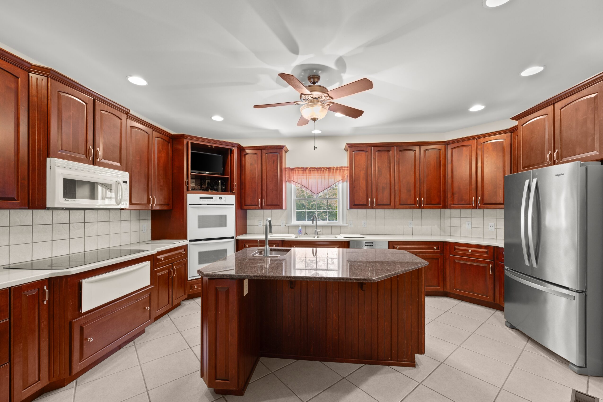 Kitchen with wooden cabinets, a central island, white appliances, tiled floor, and a ceiling fan, illuminated by ceiling lights and natural light.