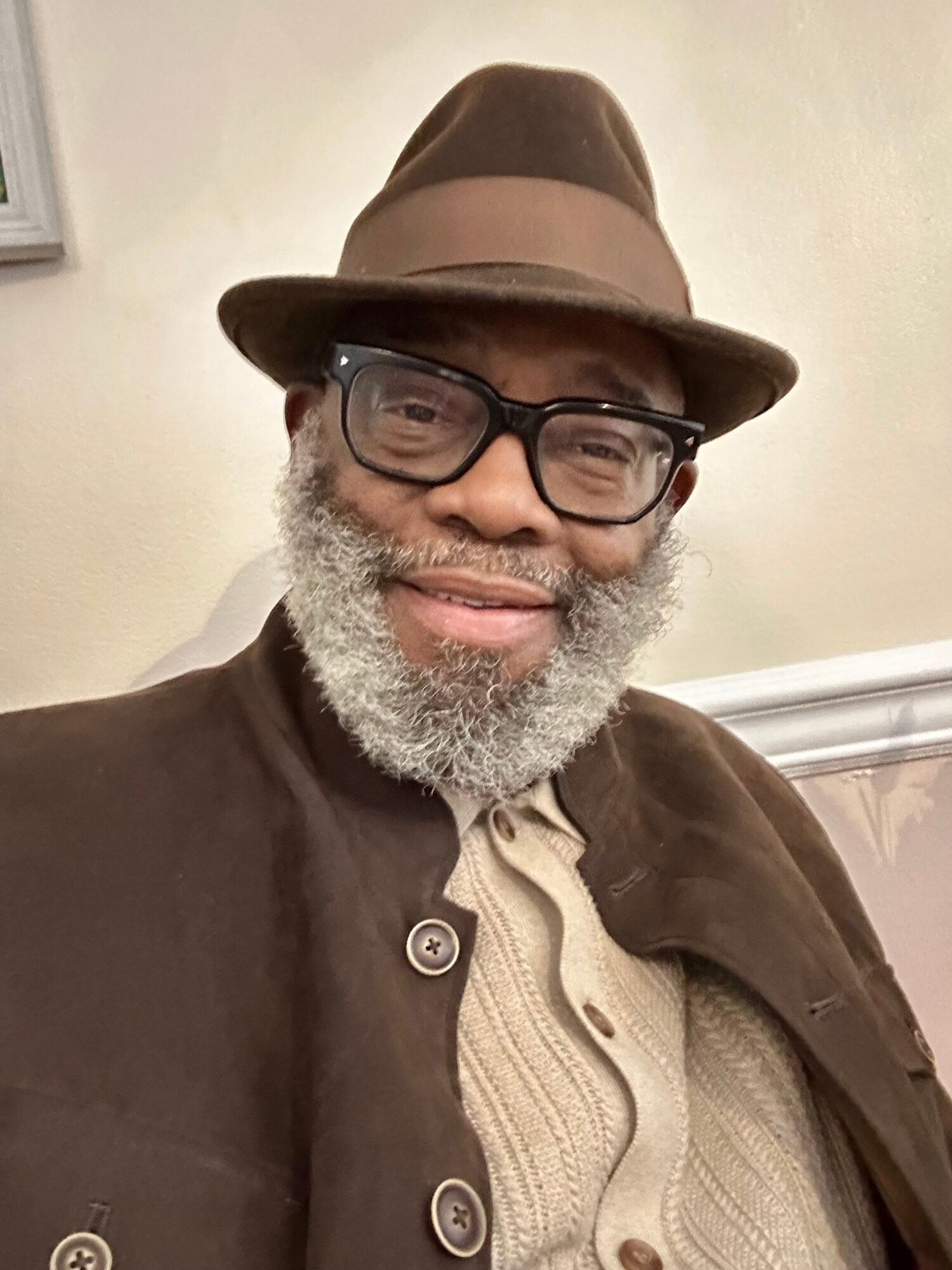 Smiling man with glasses, gray beard, wearing a brown hat and coat, beige sweater, indoors.