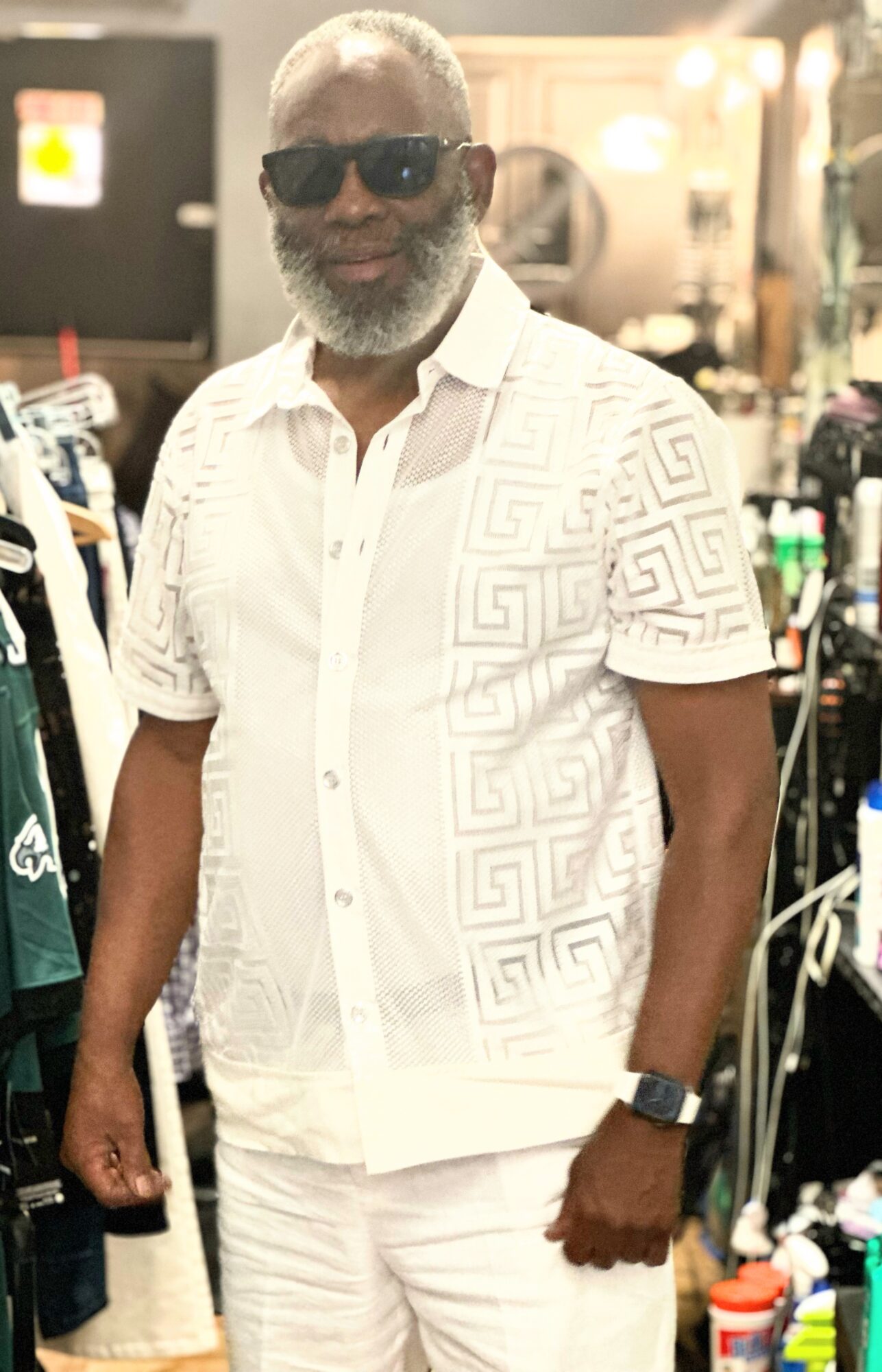 Man with gray beard and sunglasses wearing a white patterned shirt and white pants in a store.