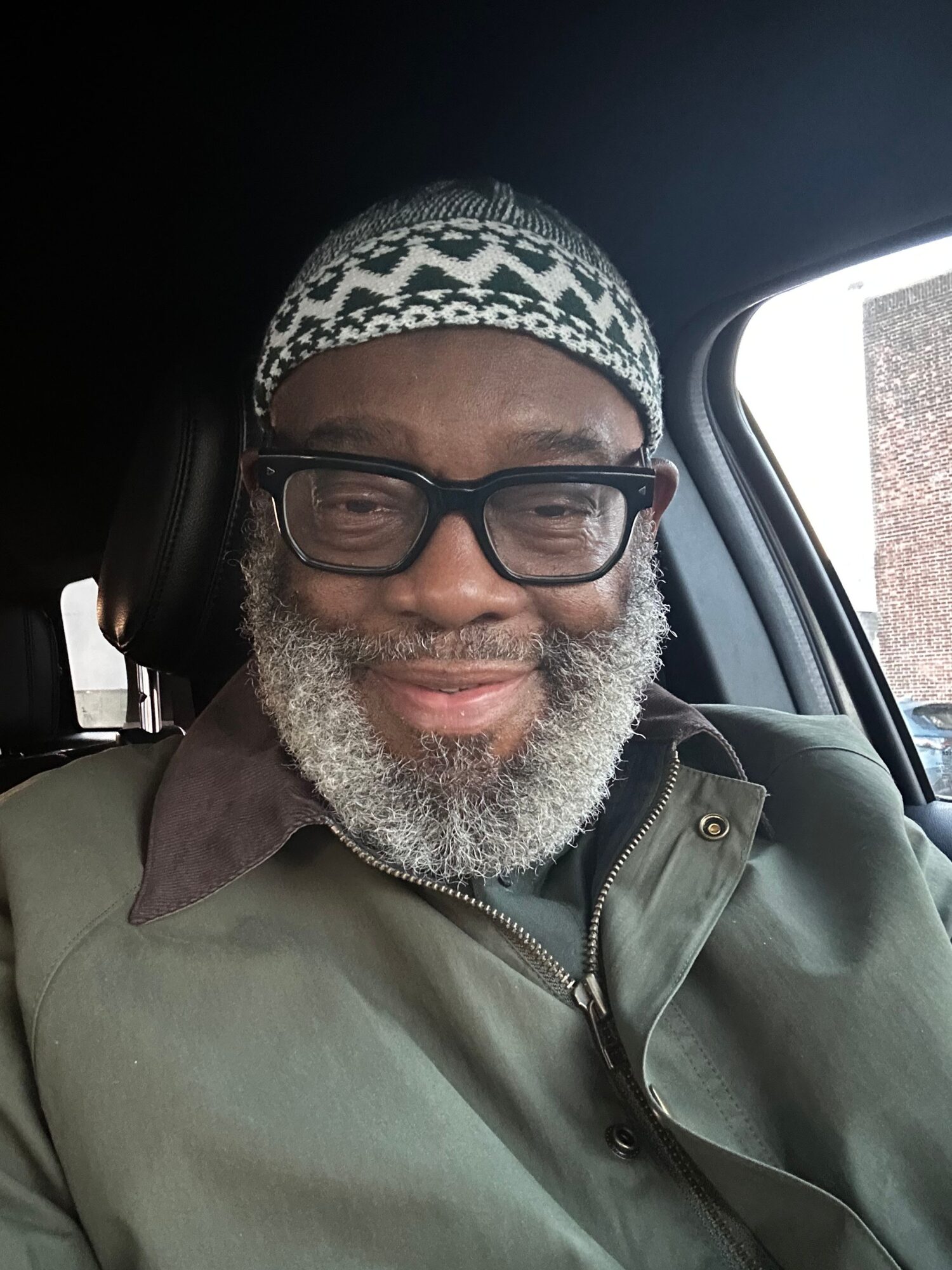 Man with glasses, gray beard, wearing a patterned hat and green jacket, sitting in vehicle, smiling.