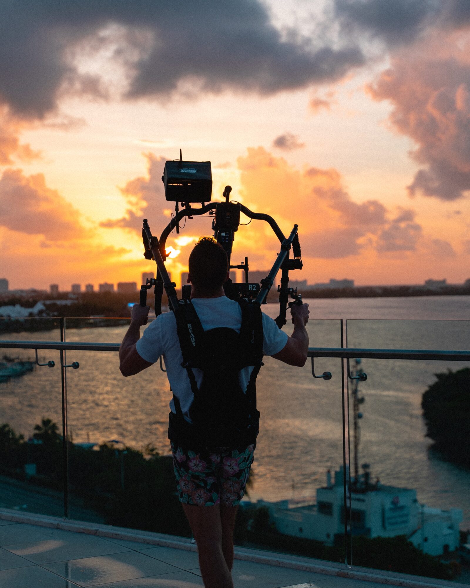 Person with a backpack holding a drone controller, facing a sunset over water with clouds and city skyline.