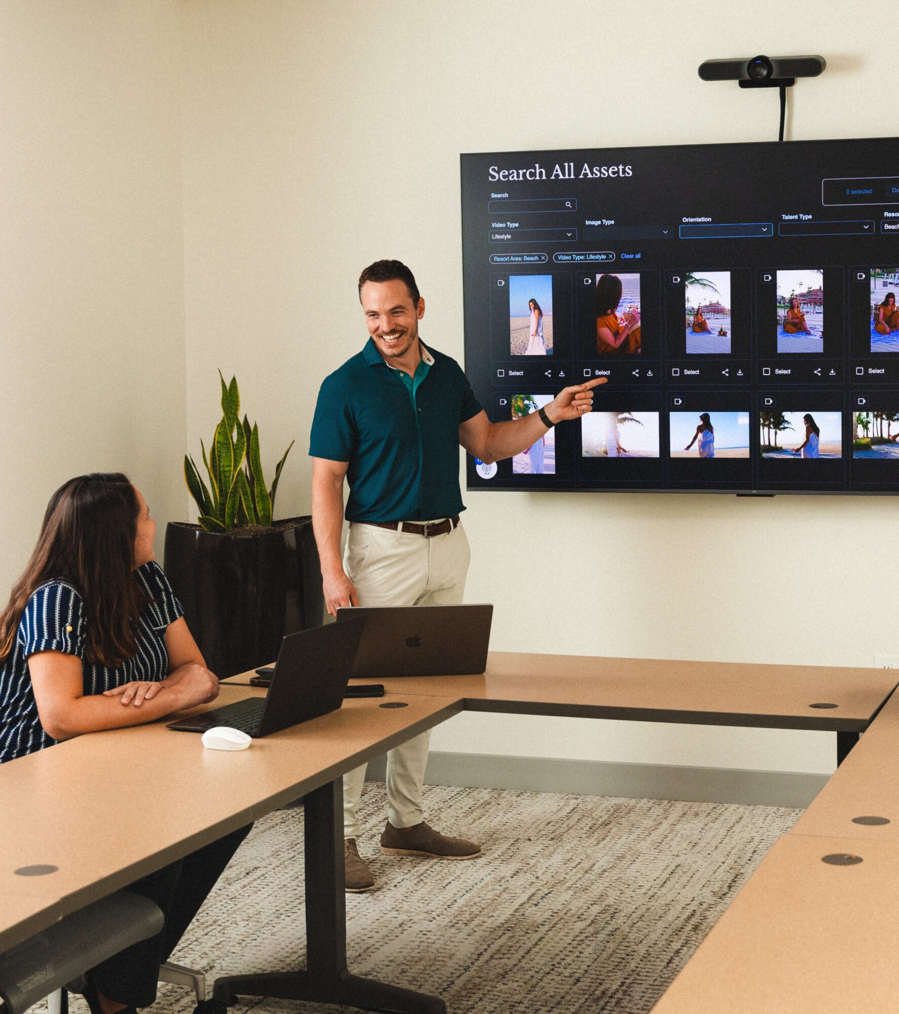 Man presenting in front of a large screen with images, two women seated at a conference table, laptop open, in a meeting room.