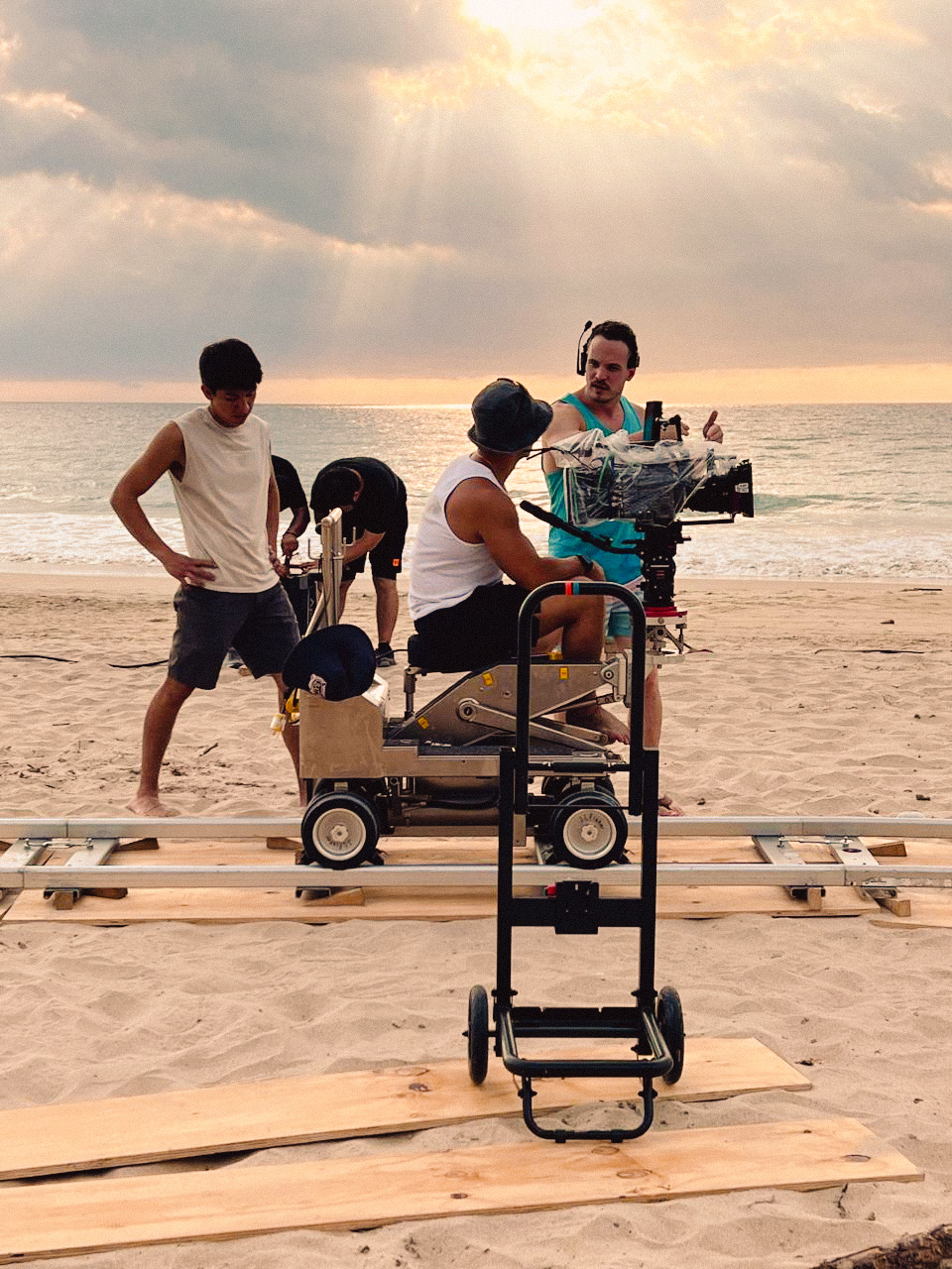 Group of people on a beach with a cart and equipment, sunlight shining through clouds, ocean in background.