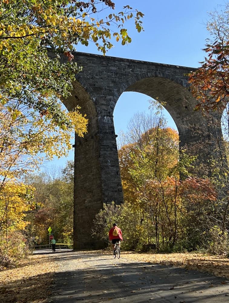 Stone arch bridge with trees and people biking underneath, clear blue sky, autumn foliage.