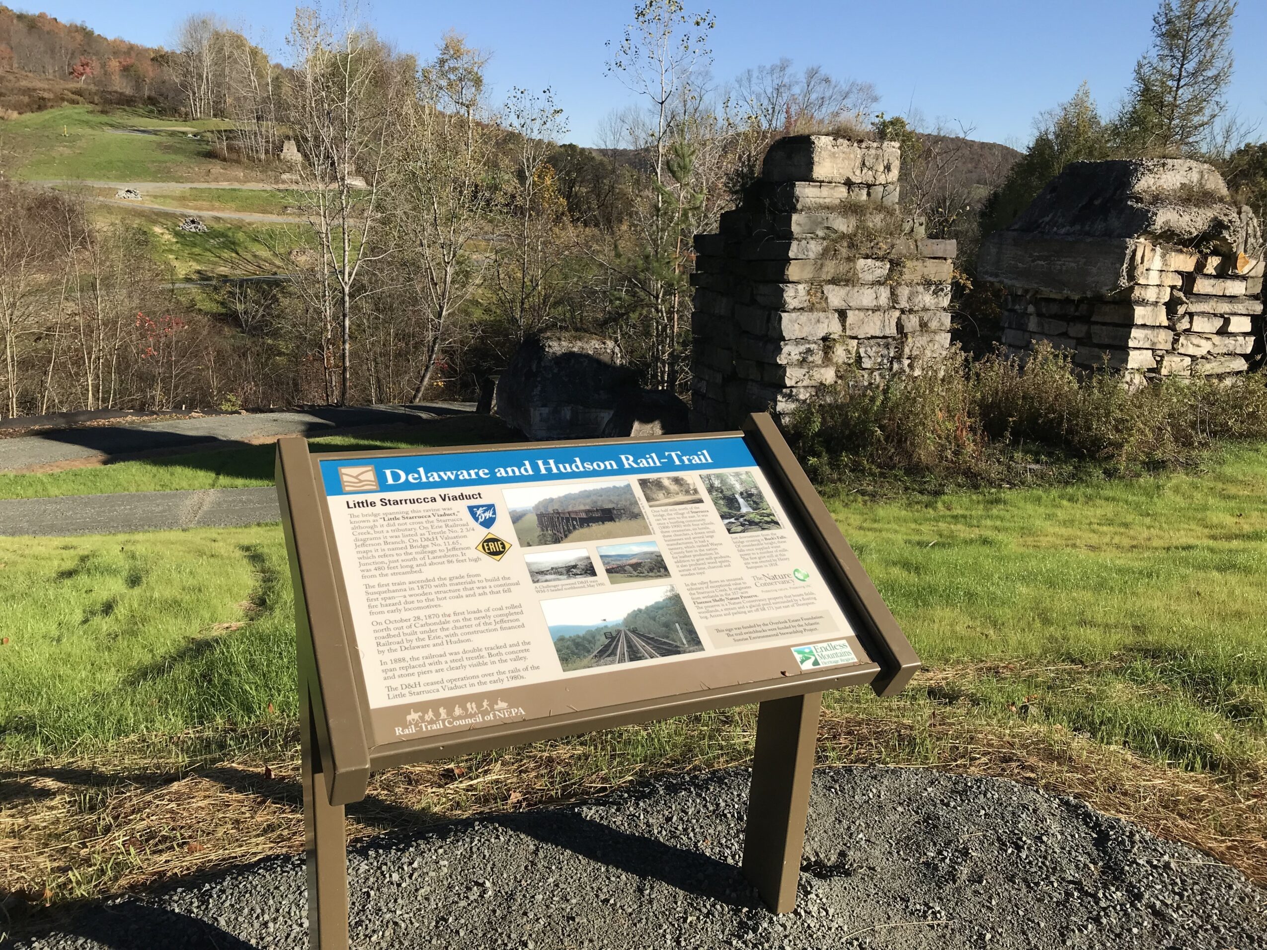 Information board titled 'Delaware and Mahanoy Rail-Trail' with text and images, set in a grassy outdoor area with ruins and trees in the background.