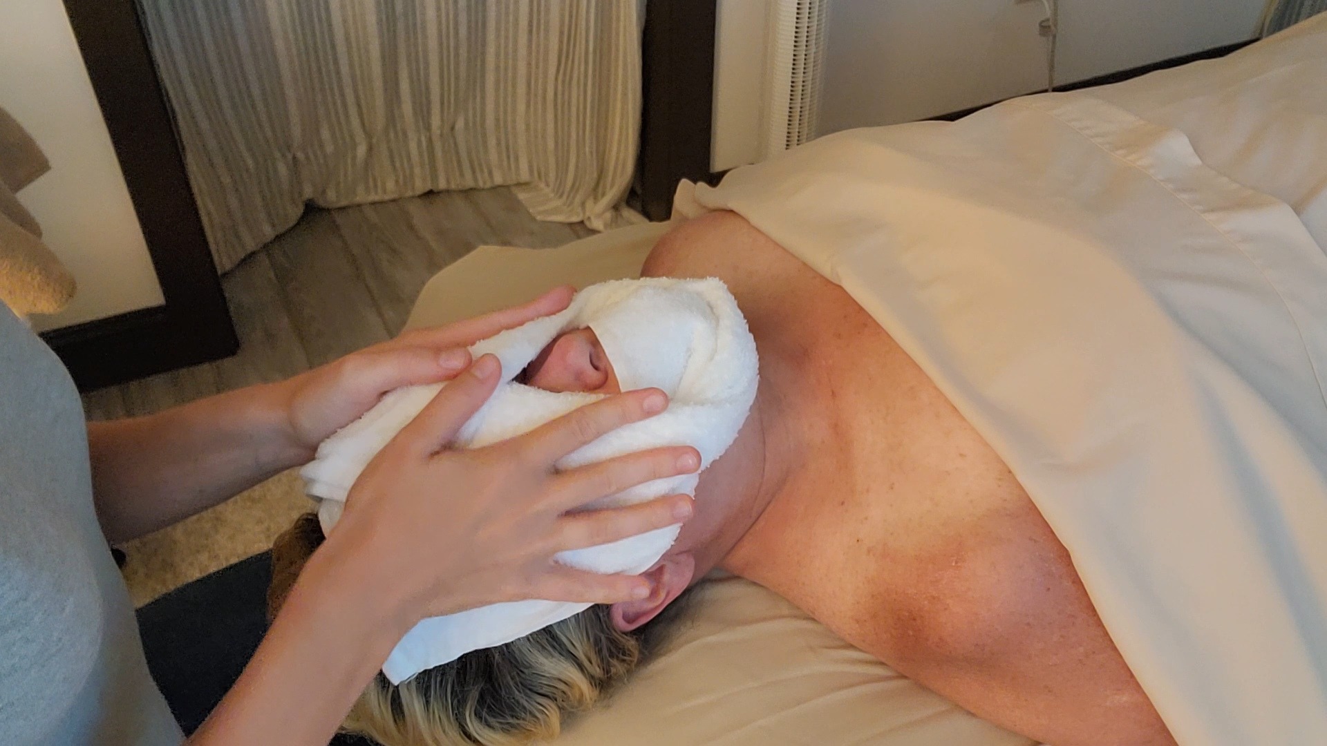 Person holding a towel or cloth over a person's face on a bed, with part of their back visible.