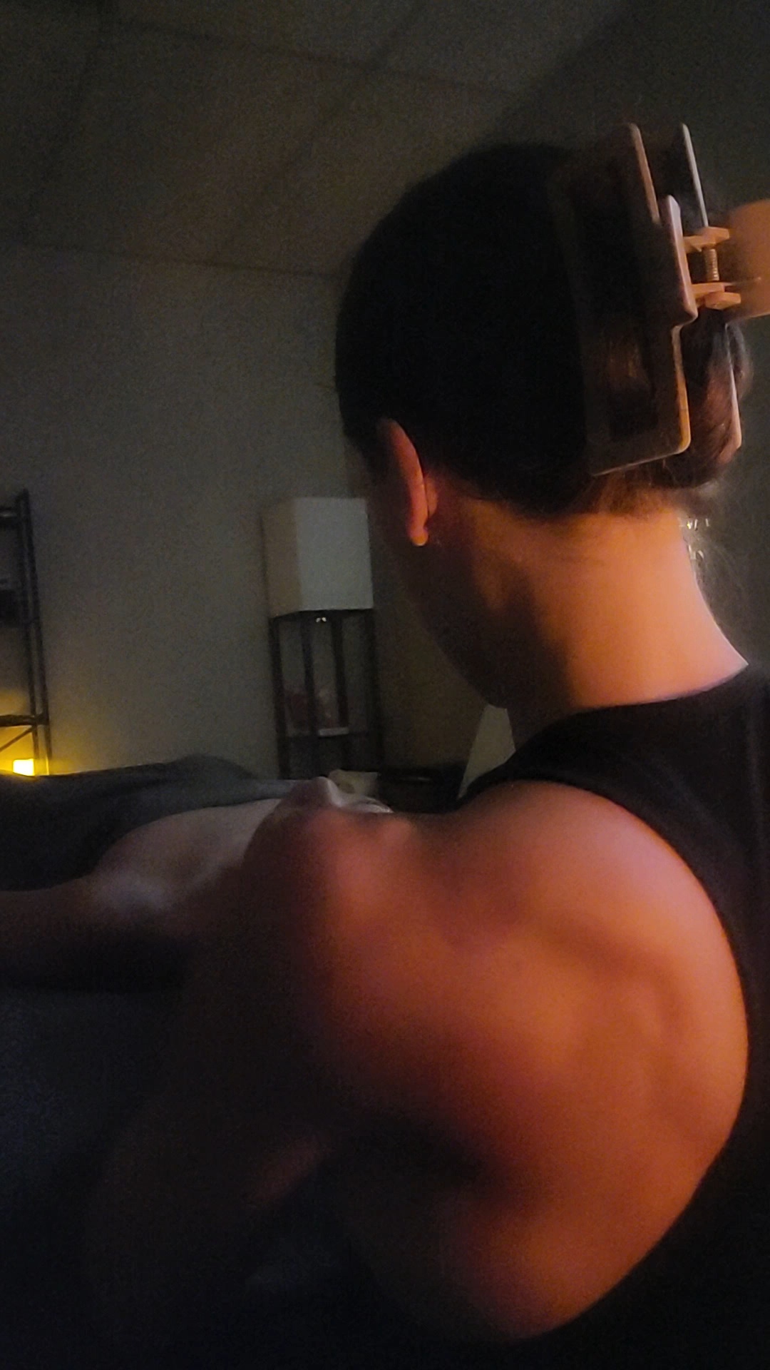 Person with short hair and a hair clip, showing their shoulder and back muscles, in a dimly lit room.