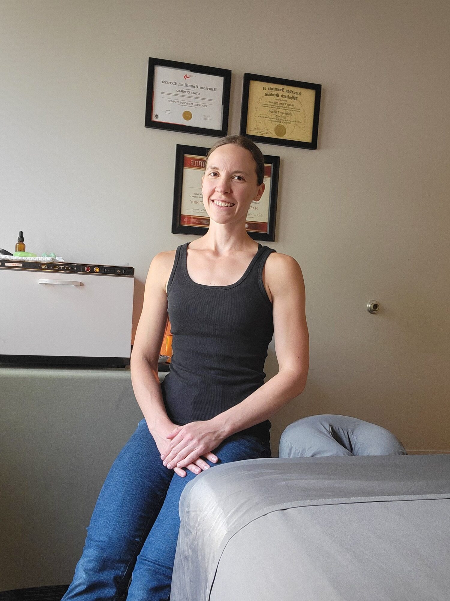 Woman standing in a room with framed certificates on the wall, wearing a black tank top and blue jeans.