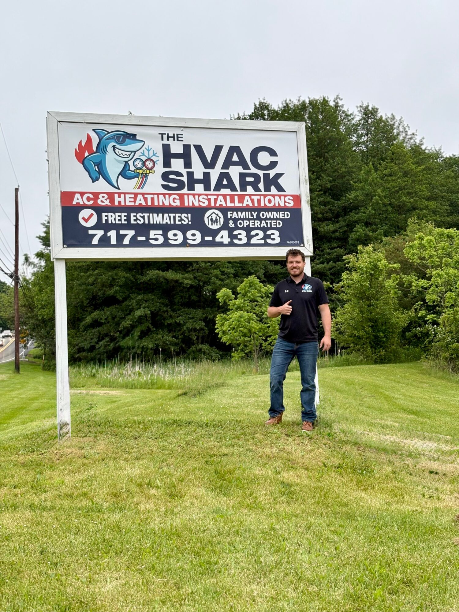 Man standing on grass next to HVAC shark billboard with trees in background.
