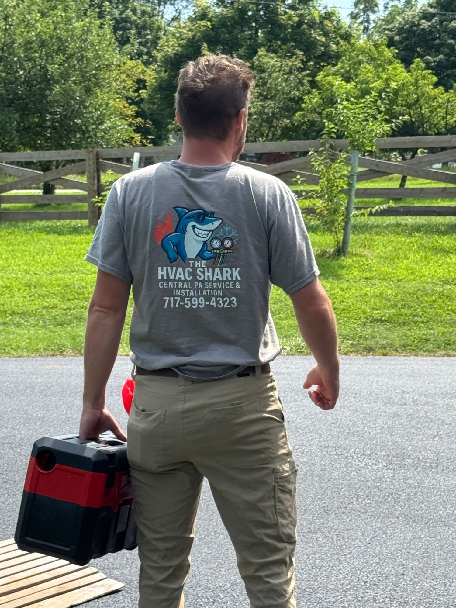 Young man standing outdoors on a paved path, holding a black and red toolbox, with trees and a wooden fence in the background.