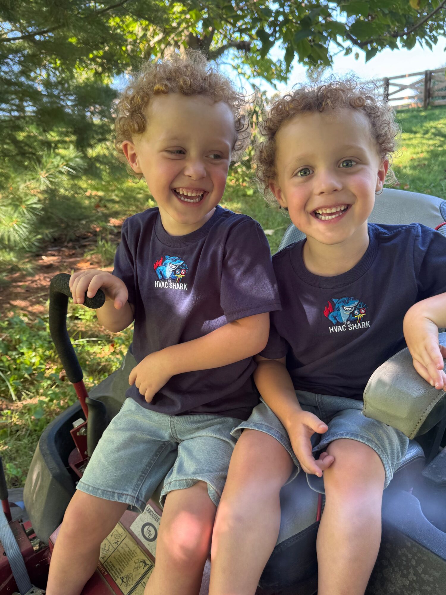Two children sitting outdoors on a sunny day, smiling, with trees in the background.