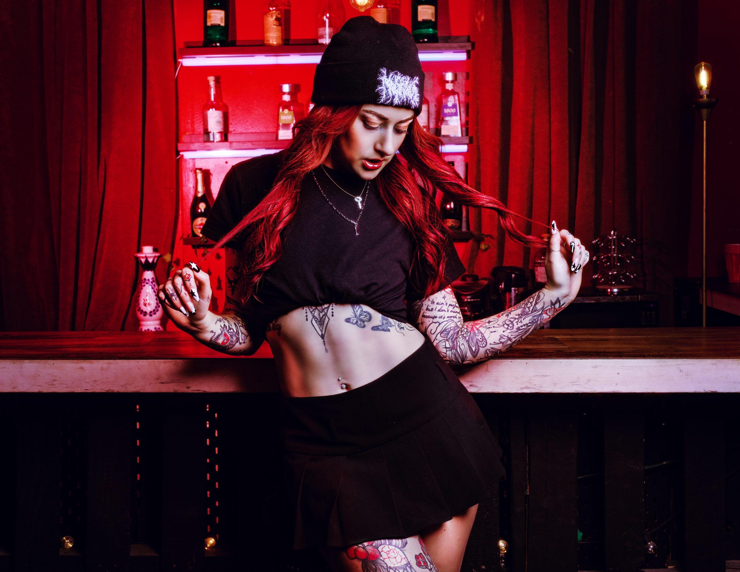 Woman with long red hair, wearing a black beanie, black crop top, and skirt, standing in front of a bar with red curtains.