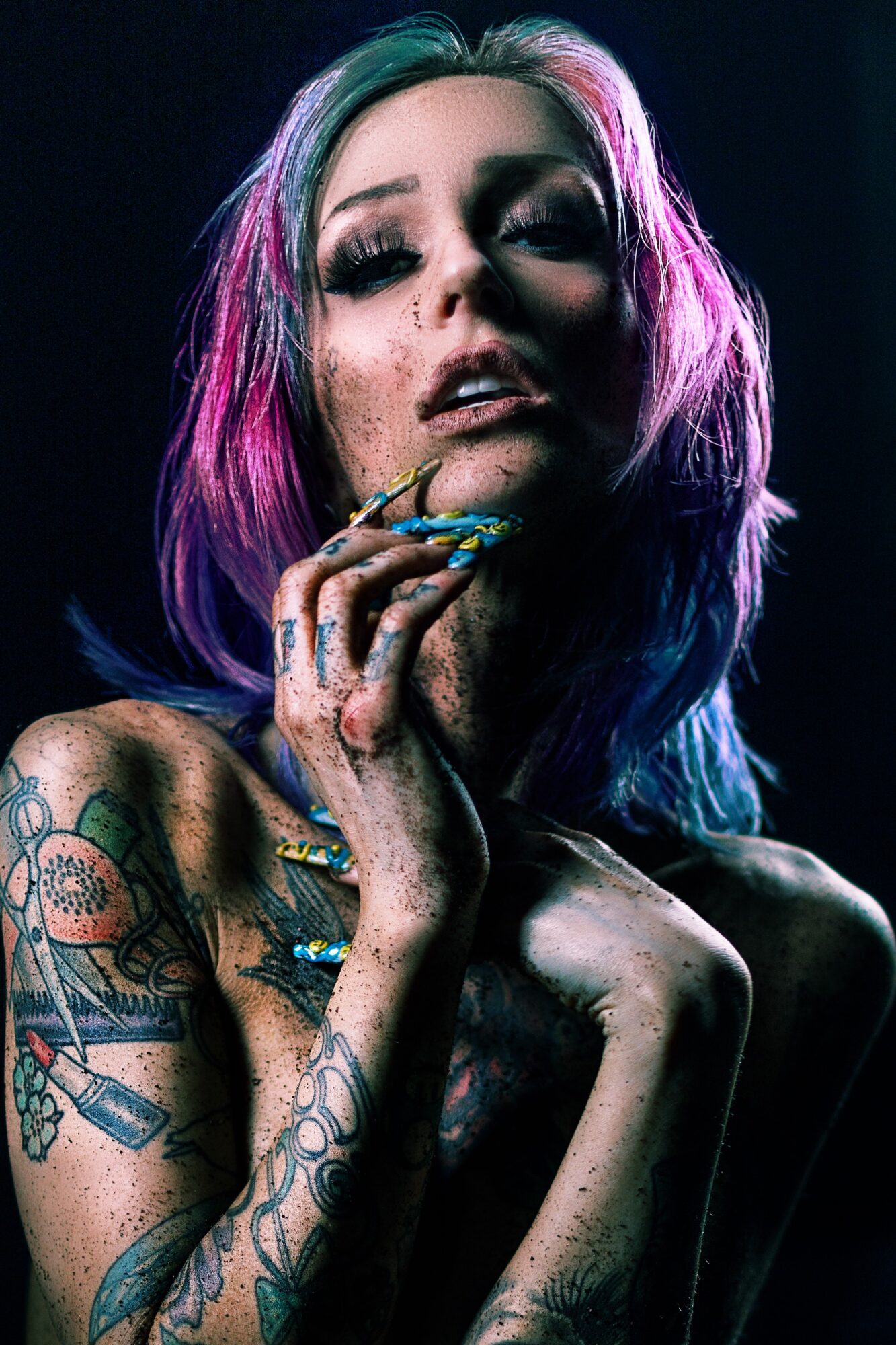 Woman with colorful hair, tattoos, and makeup, holding a cigarette, in a dark setting with dramatic lighting.