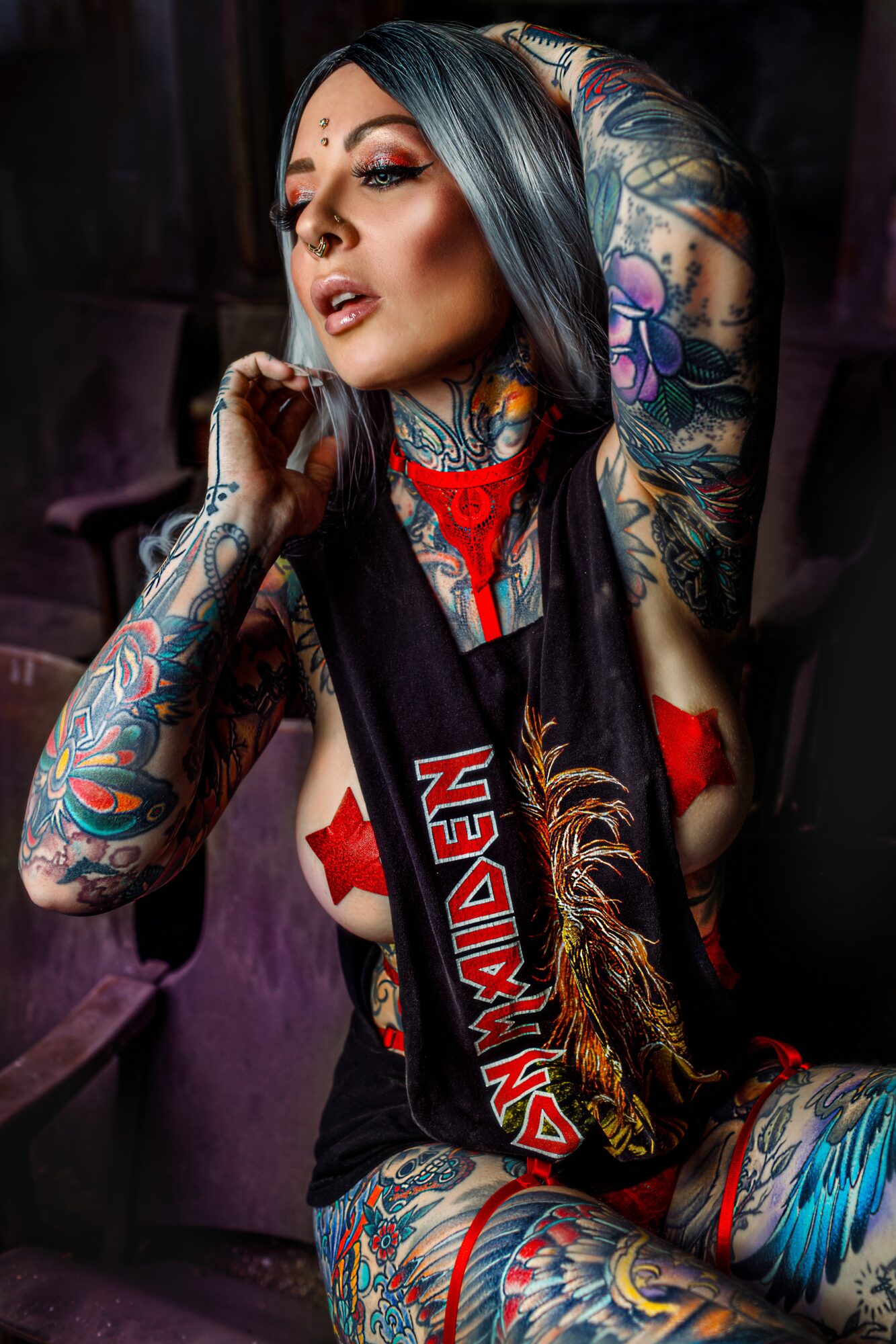 Woman with colorful tattoos, dark hair, and makeup, sitting with arms raised, wearing a black sleeveless top and red accessories.