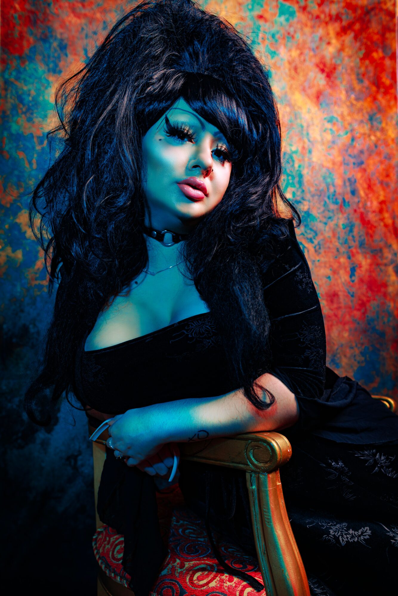Woman with dark hair and makeup, sitting on a chair against a colorful textured background.
