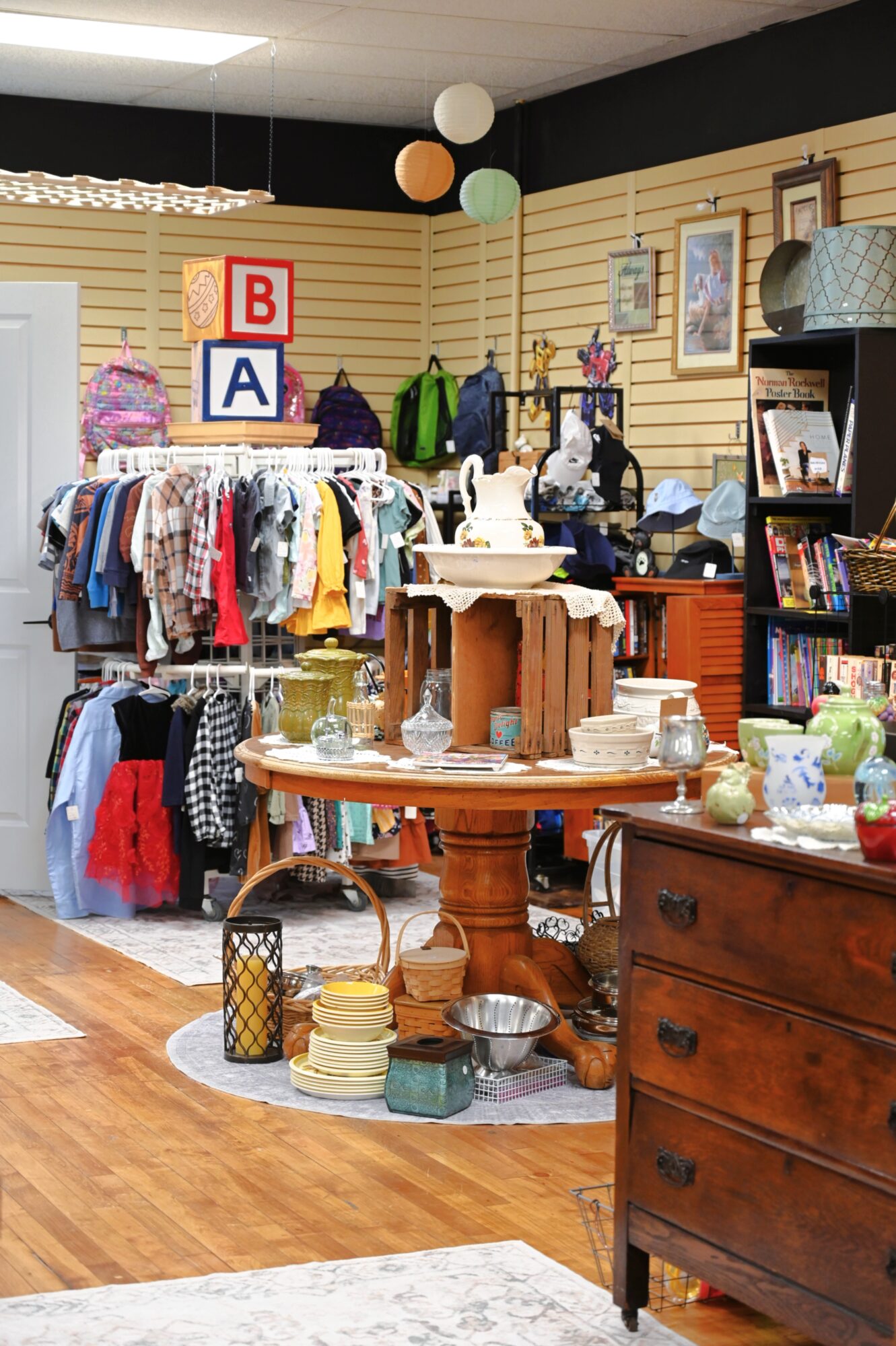 Clothing rack with various garments, a round wooden table with dishes and decor, and a wooden dresser in a store interior.