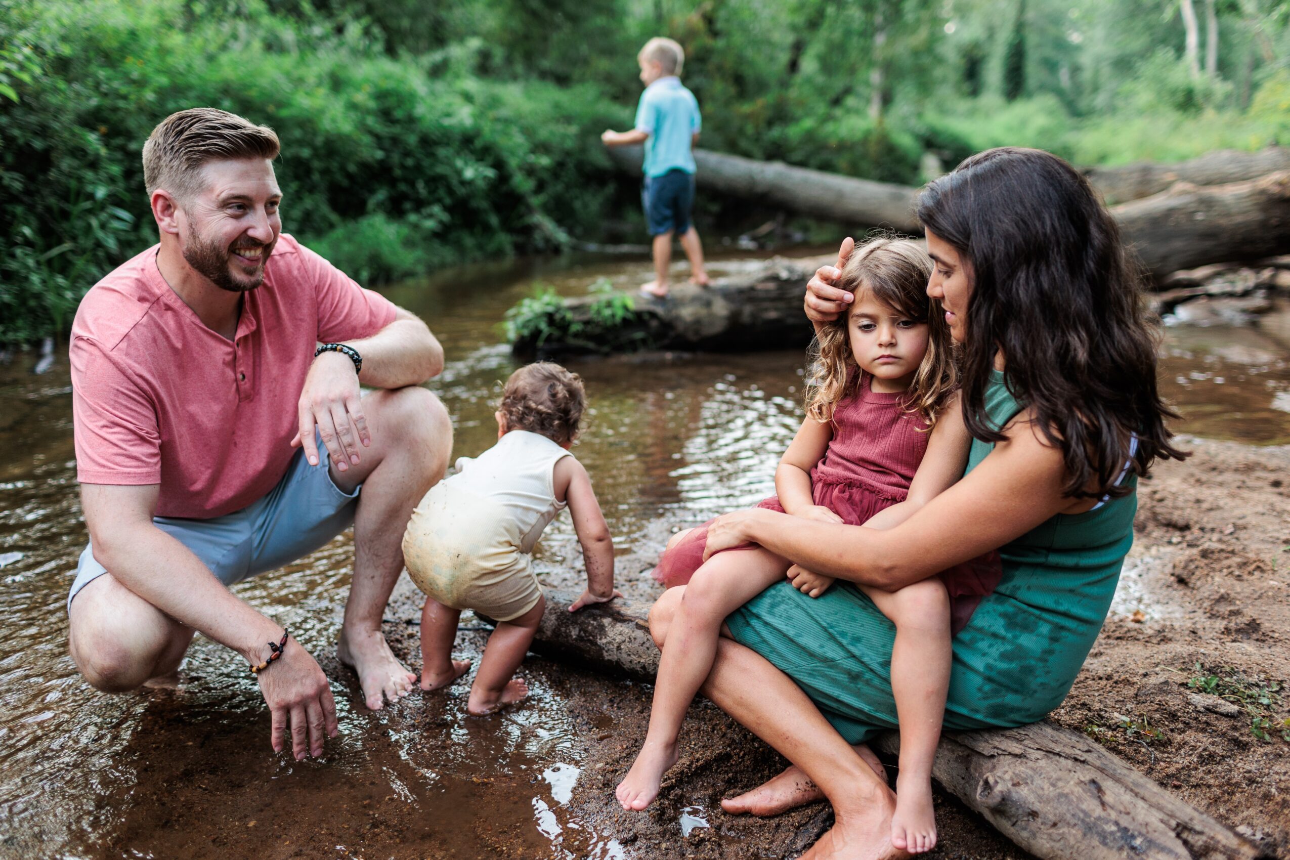 Family of four by a creek, with two children playing in the water and two adults sitting on a log, surrounded by trees.