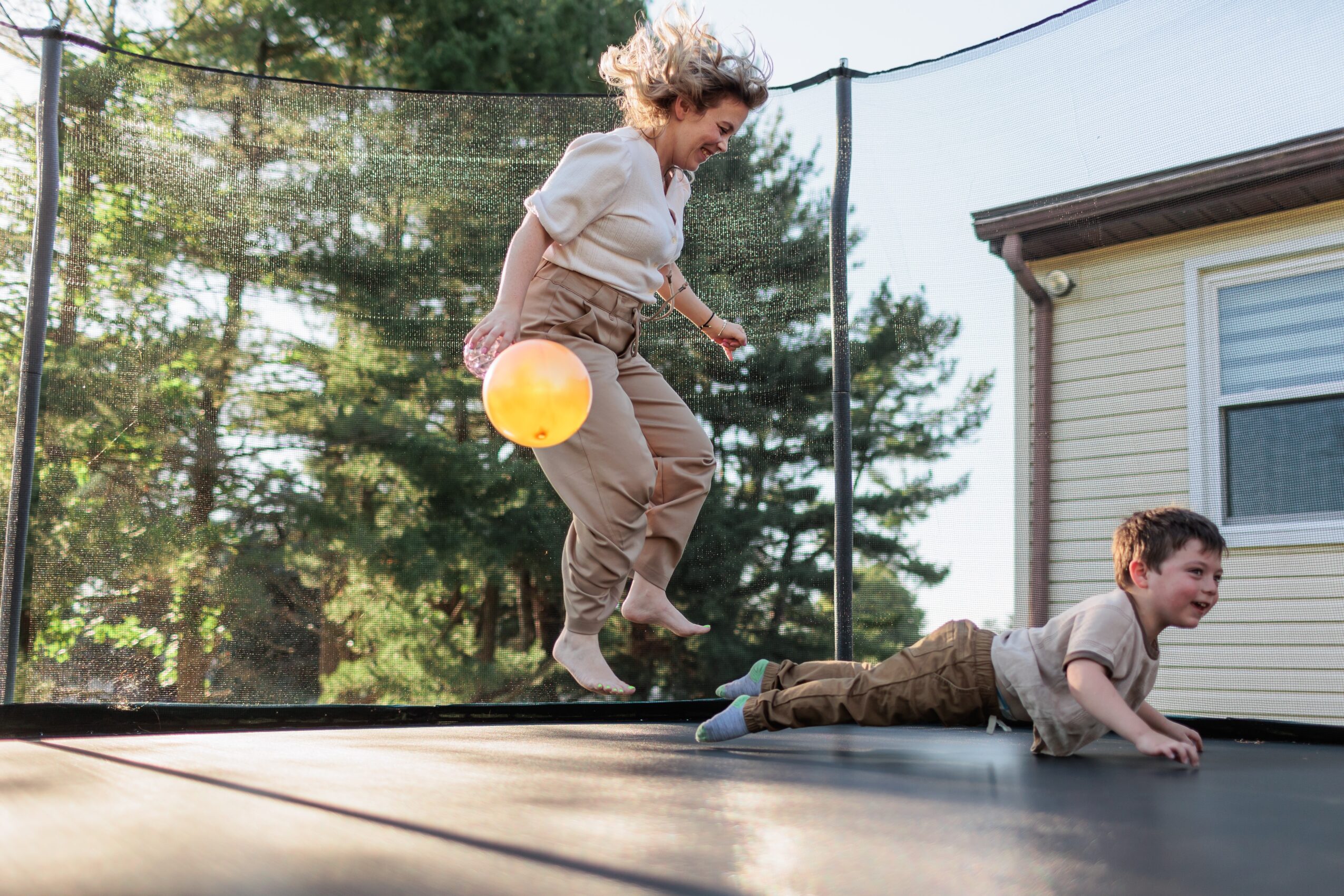 Child with curly hair jumps on a trampoline, while another child falls off onto the ground outside a house.