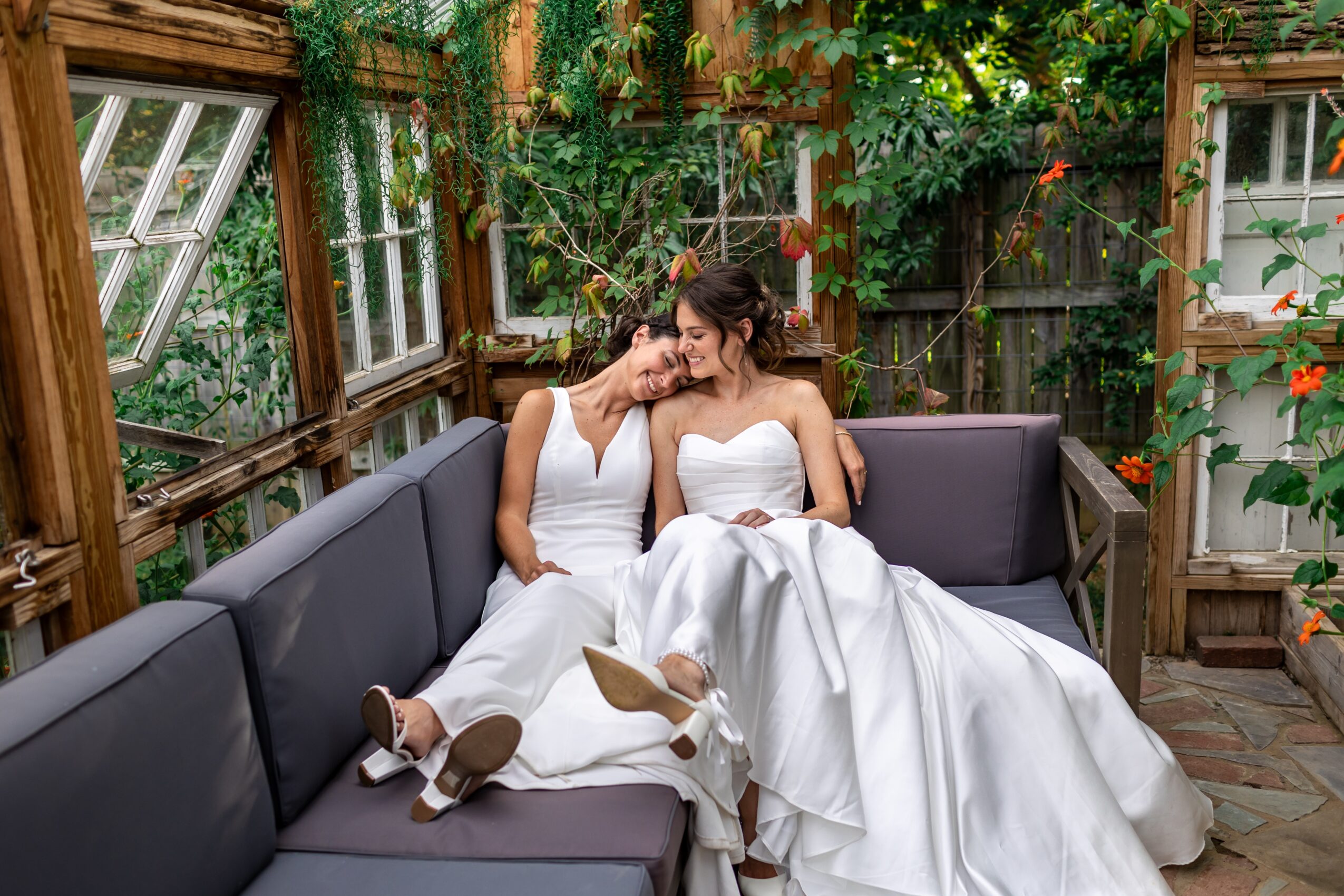 Two women in wedding dresses sit on a gray outdoor sofa in a garden with wooden structures and greenery.