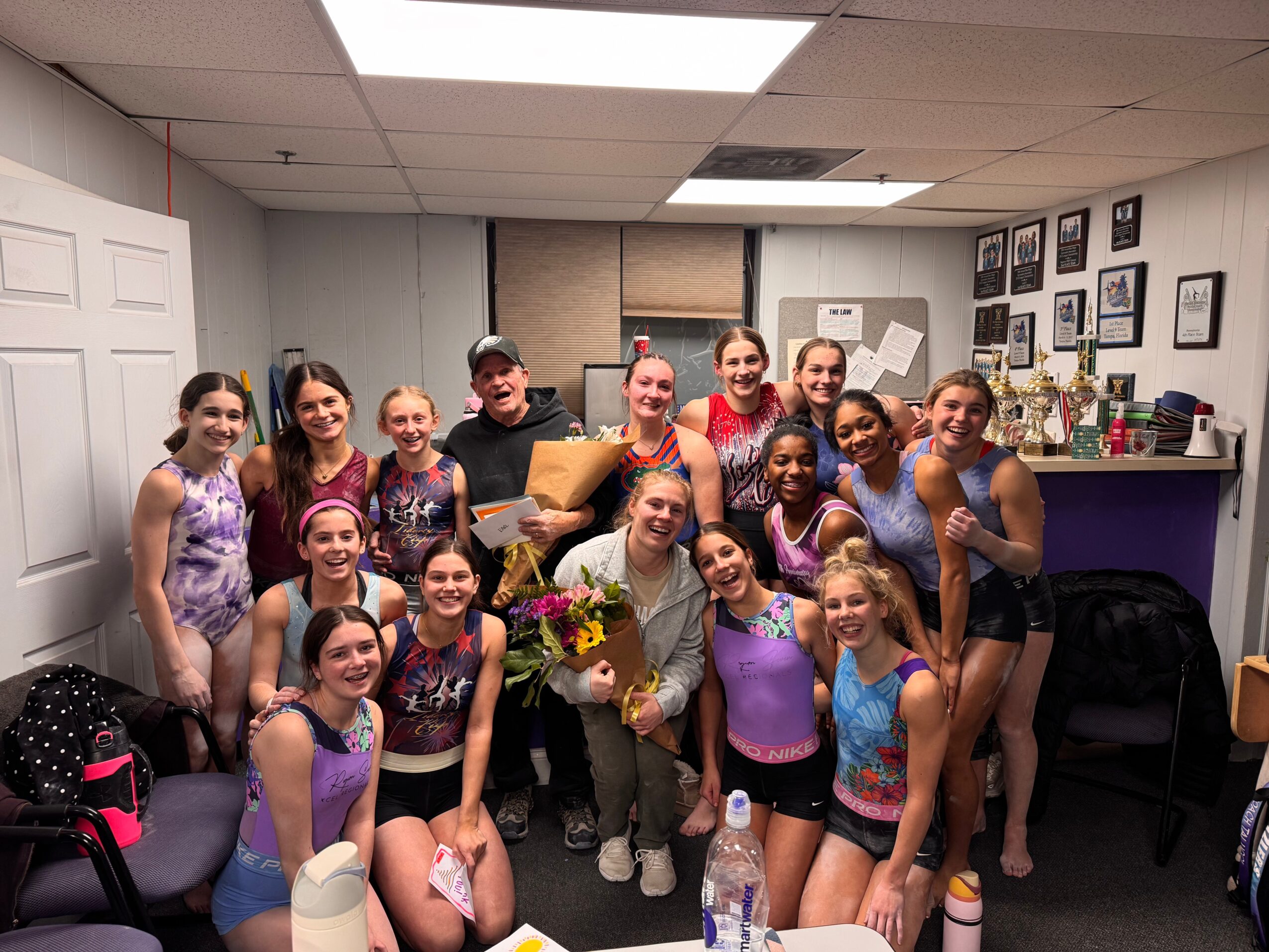 Group of young gymnasts and coaches in a room, some holding flowers, smiling for a photo.