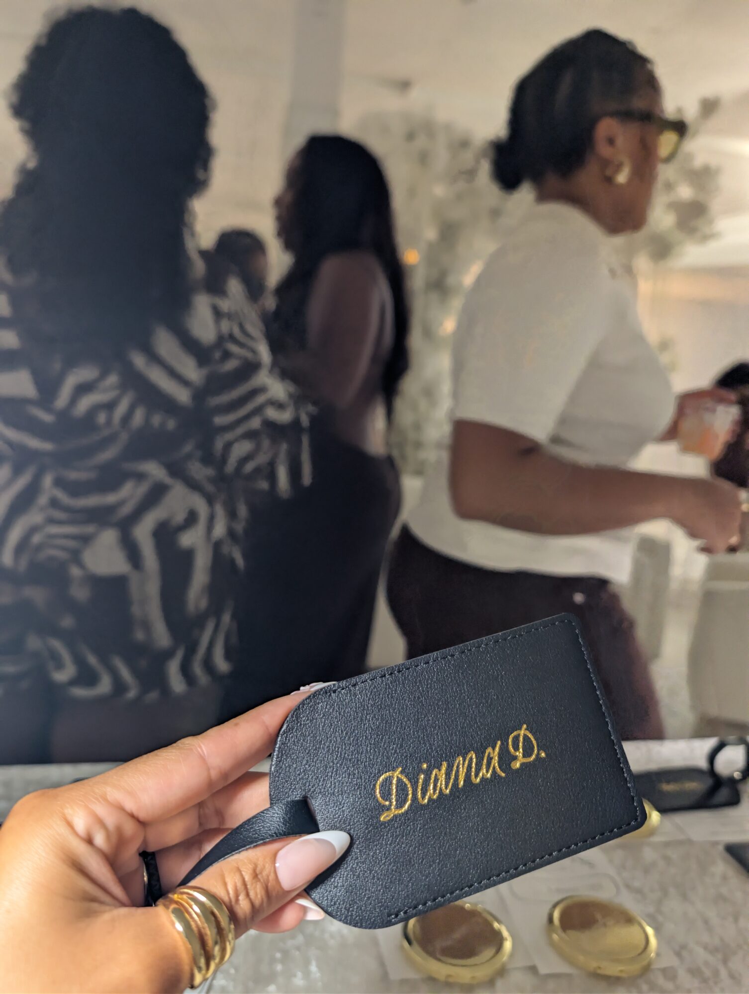 Person holding a black cardholder with gold embroidered text, with people in the background outdoors.