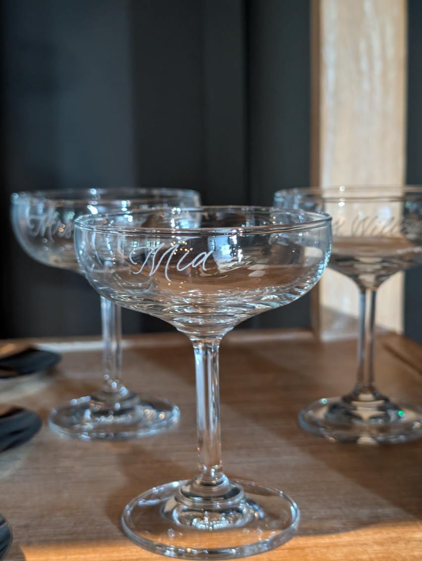 Several empty glass coupe glasses on a wooden table, with a window in the background, natural light illuminating the scene.
