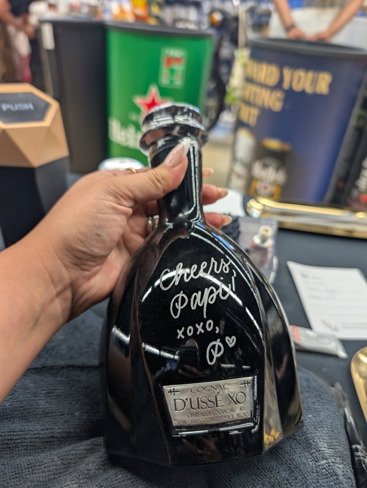 Hand holding a black bottle with handwritten-style text and symbols, in a bar or store setting.