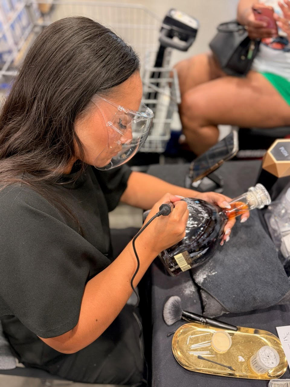 Person wearing glasses and black shirt working with a tool on a plastic bottle, with another person in the background holding a device.