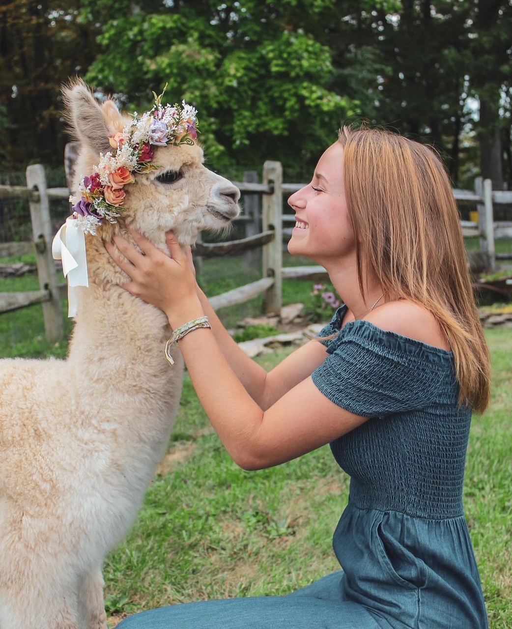 Young woman smiling at a llama with a floral crown outdoors, holding its face gently.