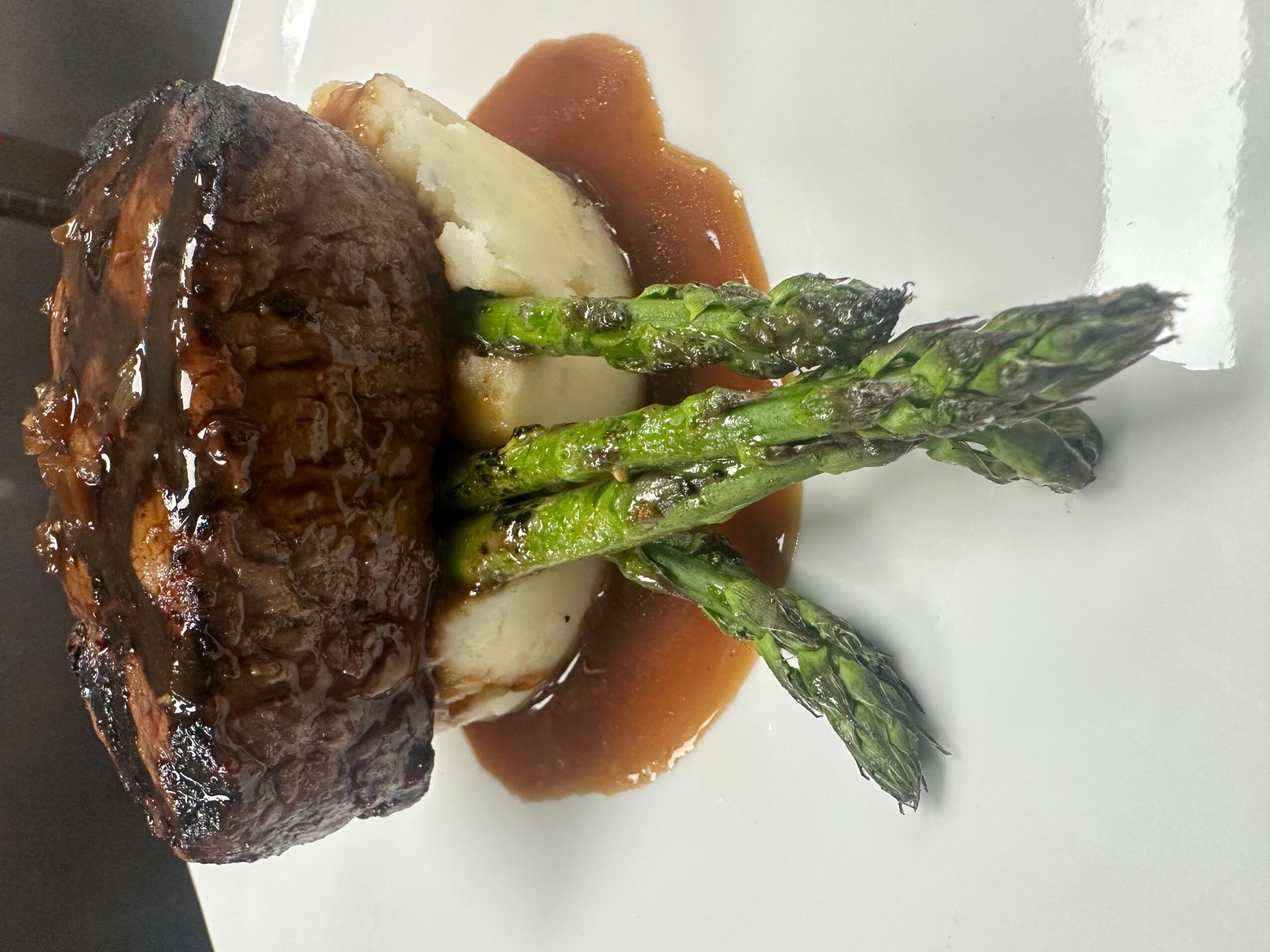 Cooked steak with asparagus and mashed potatoes on a white plate, with sauce drizzled over the steak.