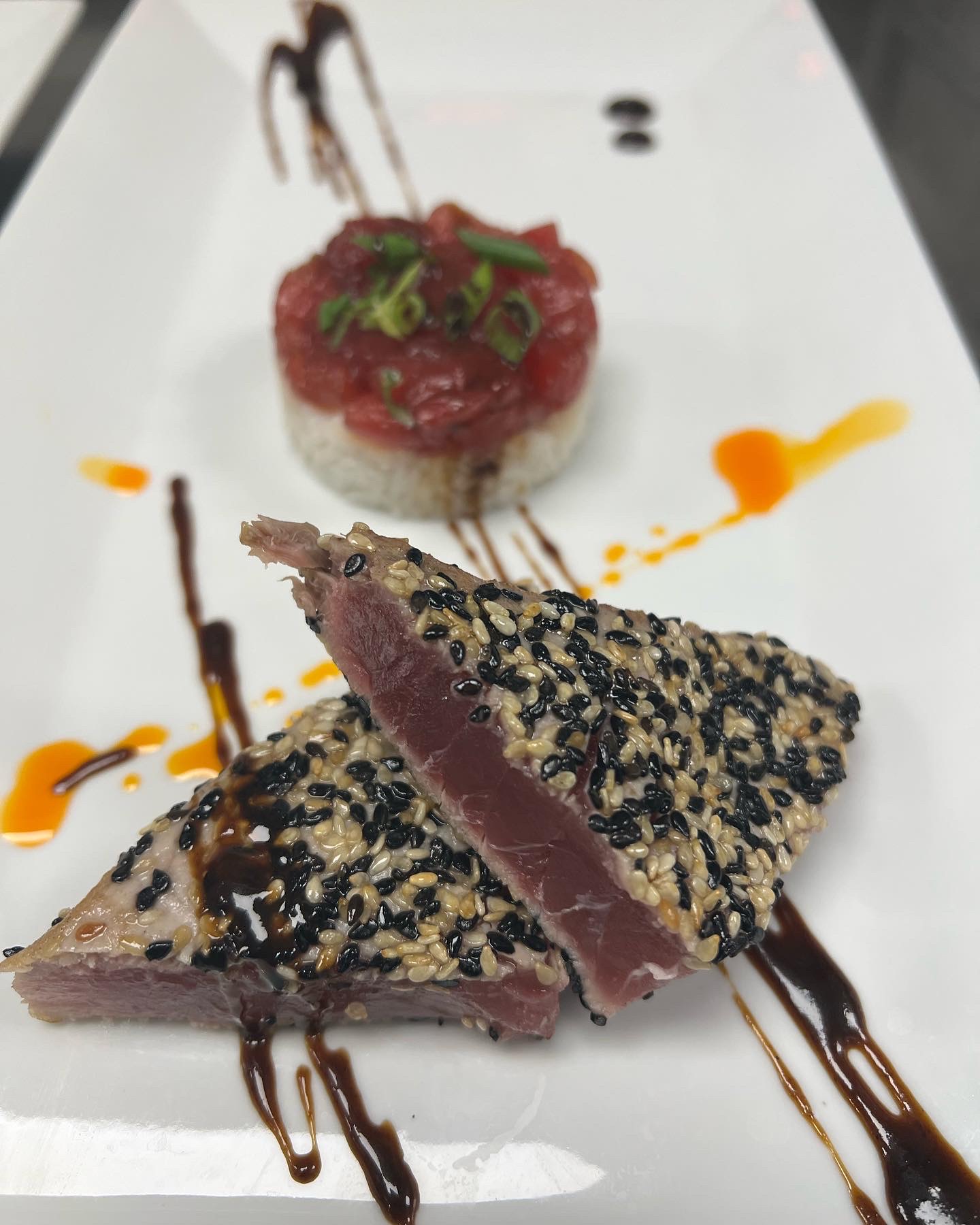 Sliced seared tuna with sesame seeds, topped with black sesame seeds, on a white plate with sauce drizzles, and a small round dish with a red topping and green garnish in the background.
