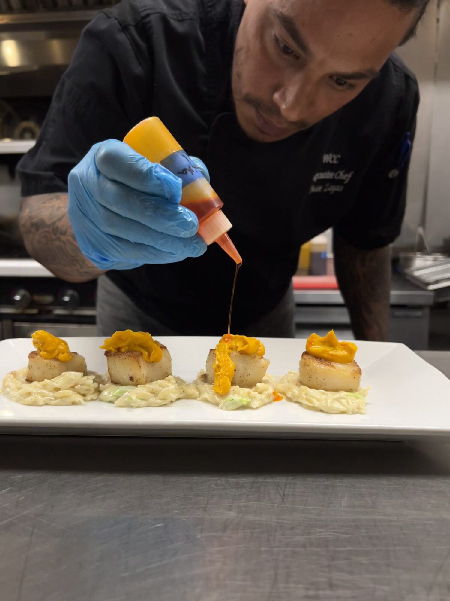 Chef decorates small appetizers with orange sauce on a white plate in a kitchen, wearing blue gloves.