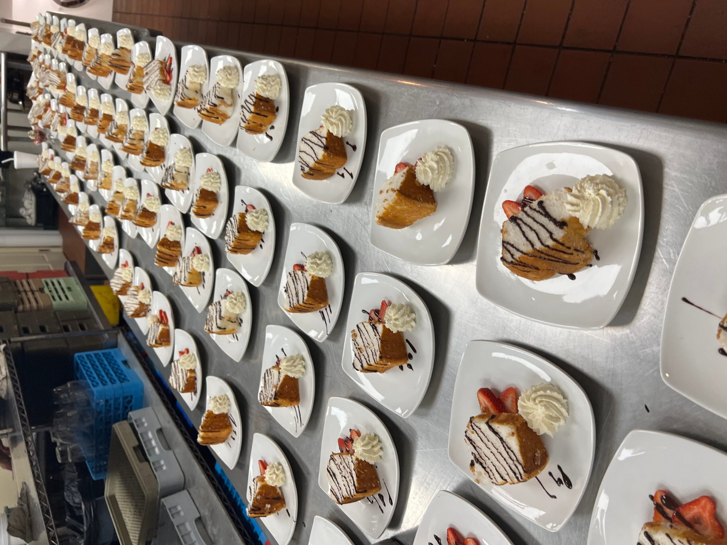 Multiple plates with slices of cake and whipped cream, arranged on a long table, viewed from the side.