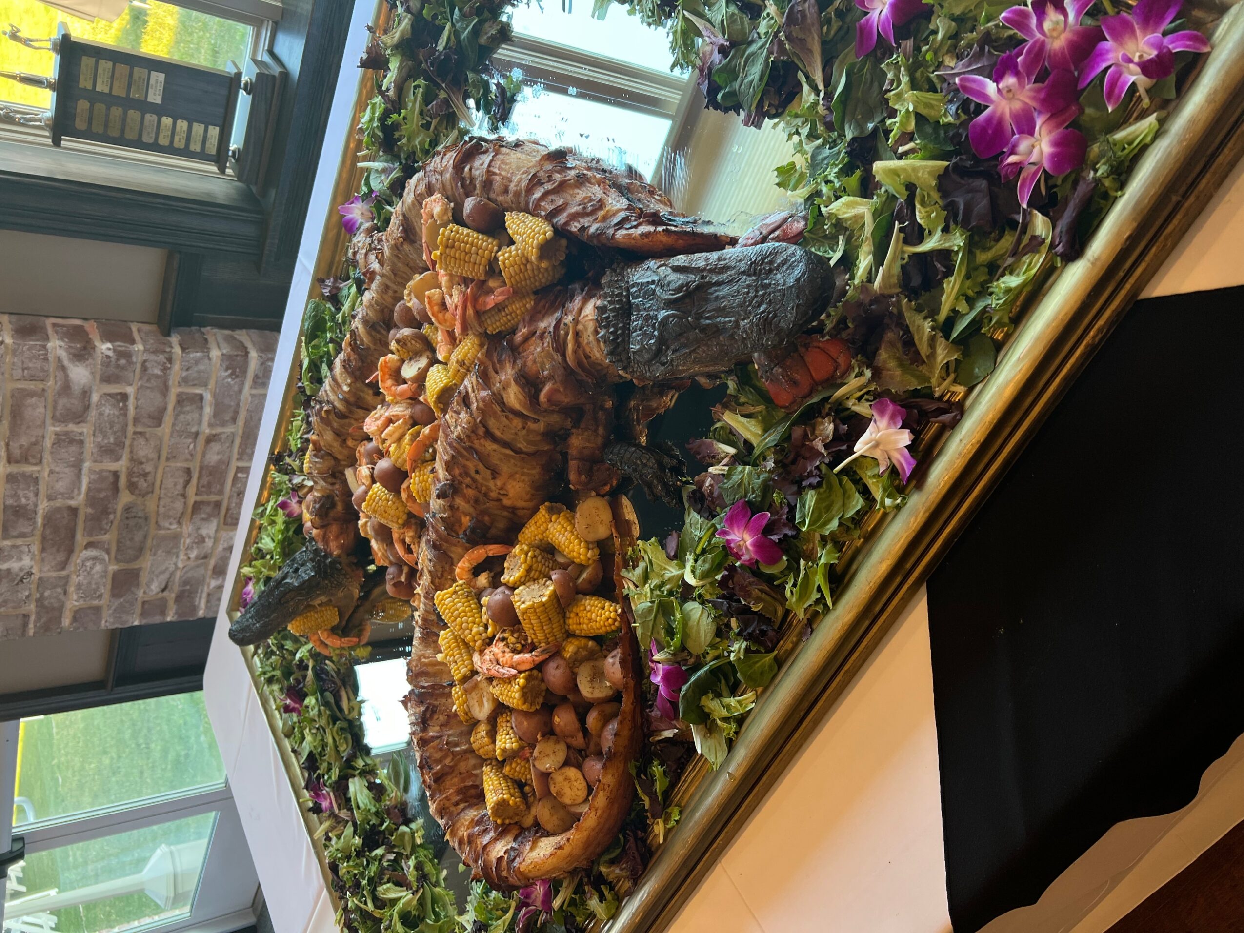 A detailed model of a dinosaur surrounded by flowers and greenery indoors, with a window and brick wall in the background.
