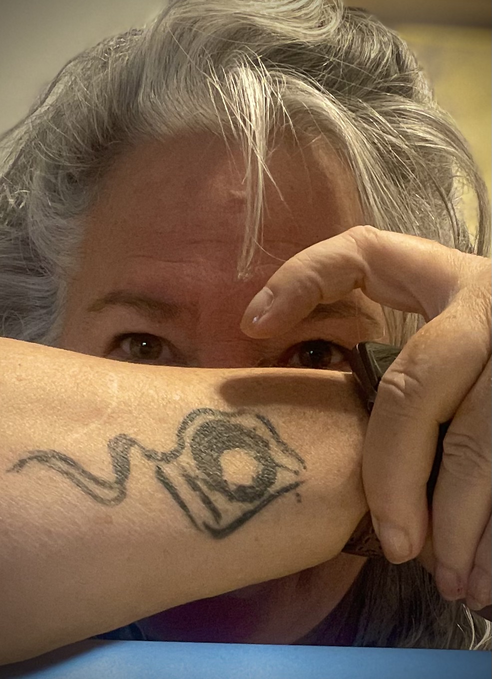 Person with gray hair partially visible, holding arm with a tattoo over their face, background blurred.