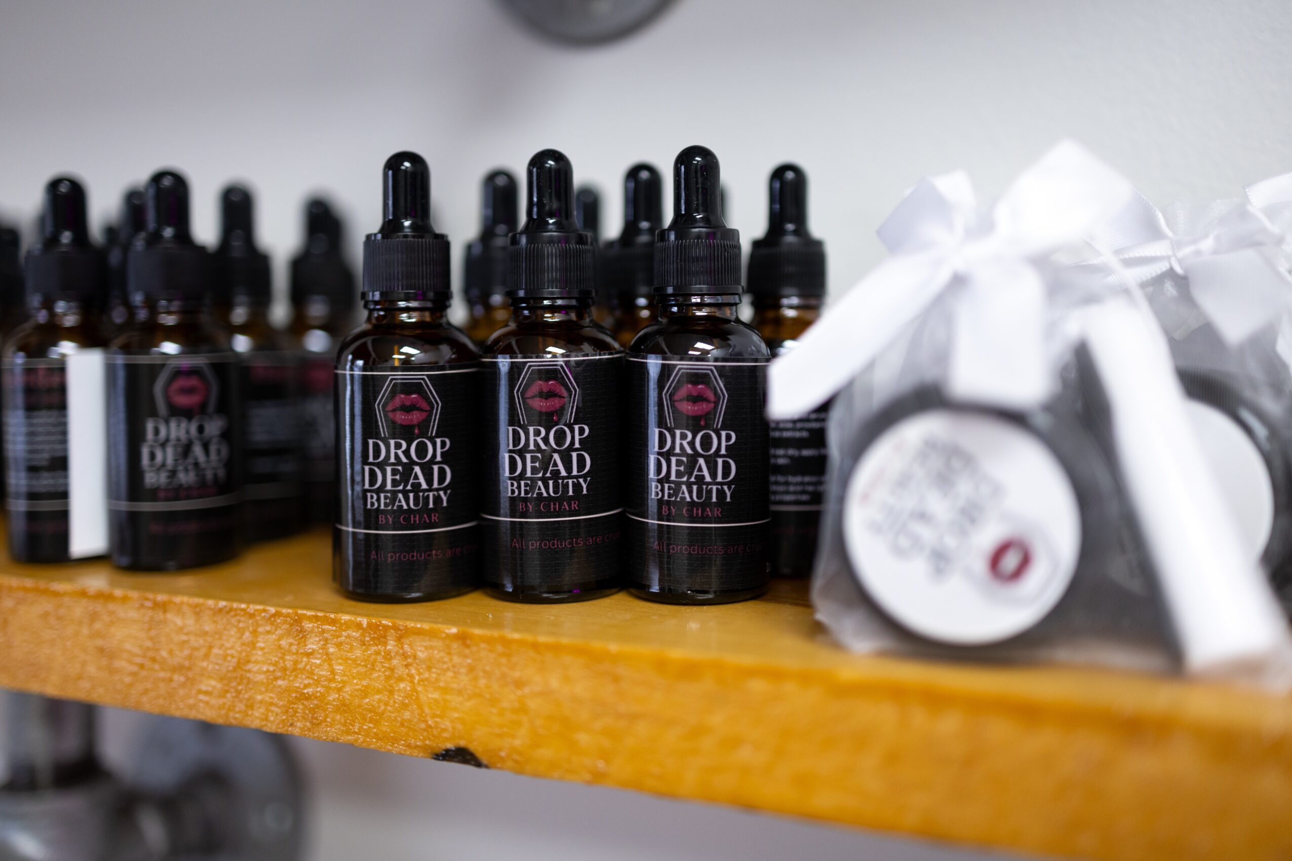 Several small bottles labeled Drop Dead Beauty on a wooden shelf, with white wrapped items nearby.