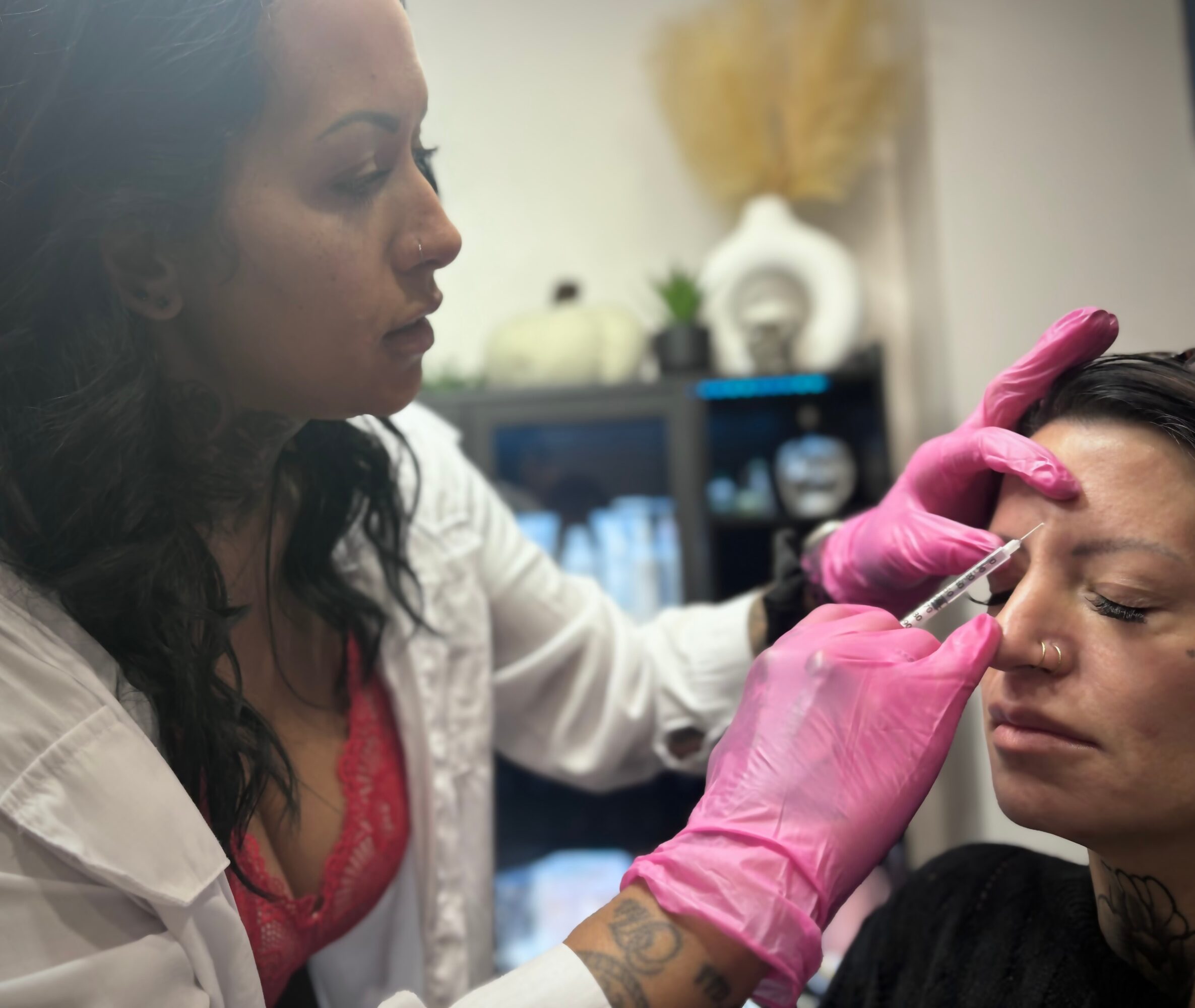 A woman in pink gloves performs a cosmetic procedure on another woman's forehead in a clinical setting.